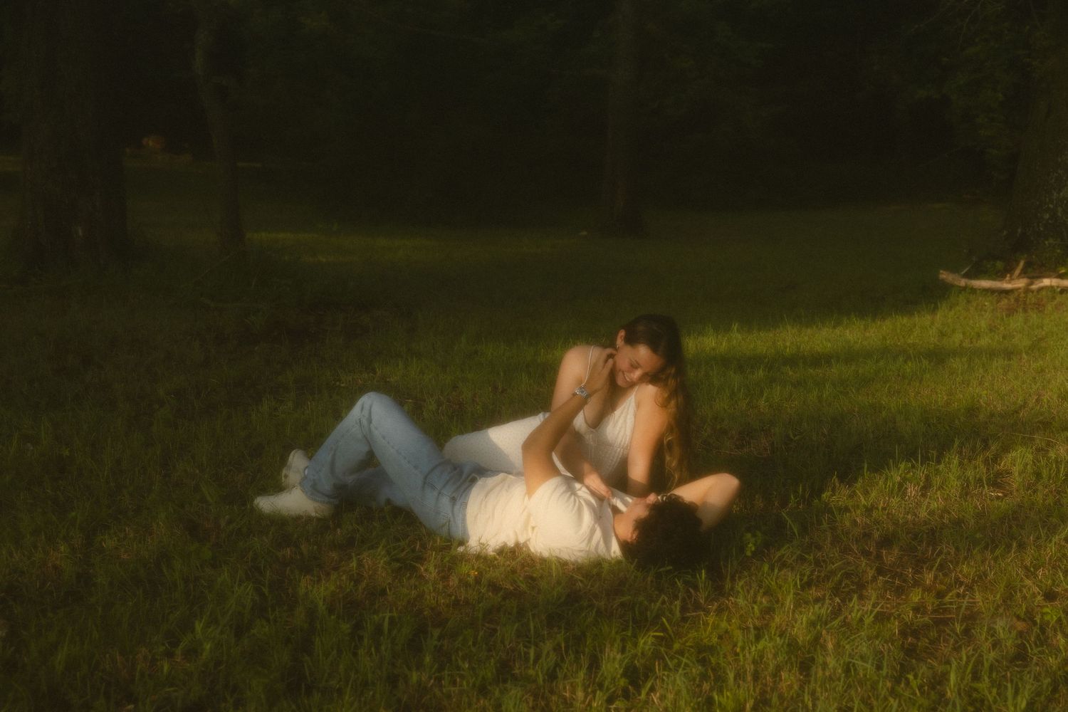 Couple lying together on lush green grass in warm golden sunlight, sharing a romantic moment outdoors.