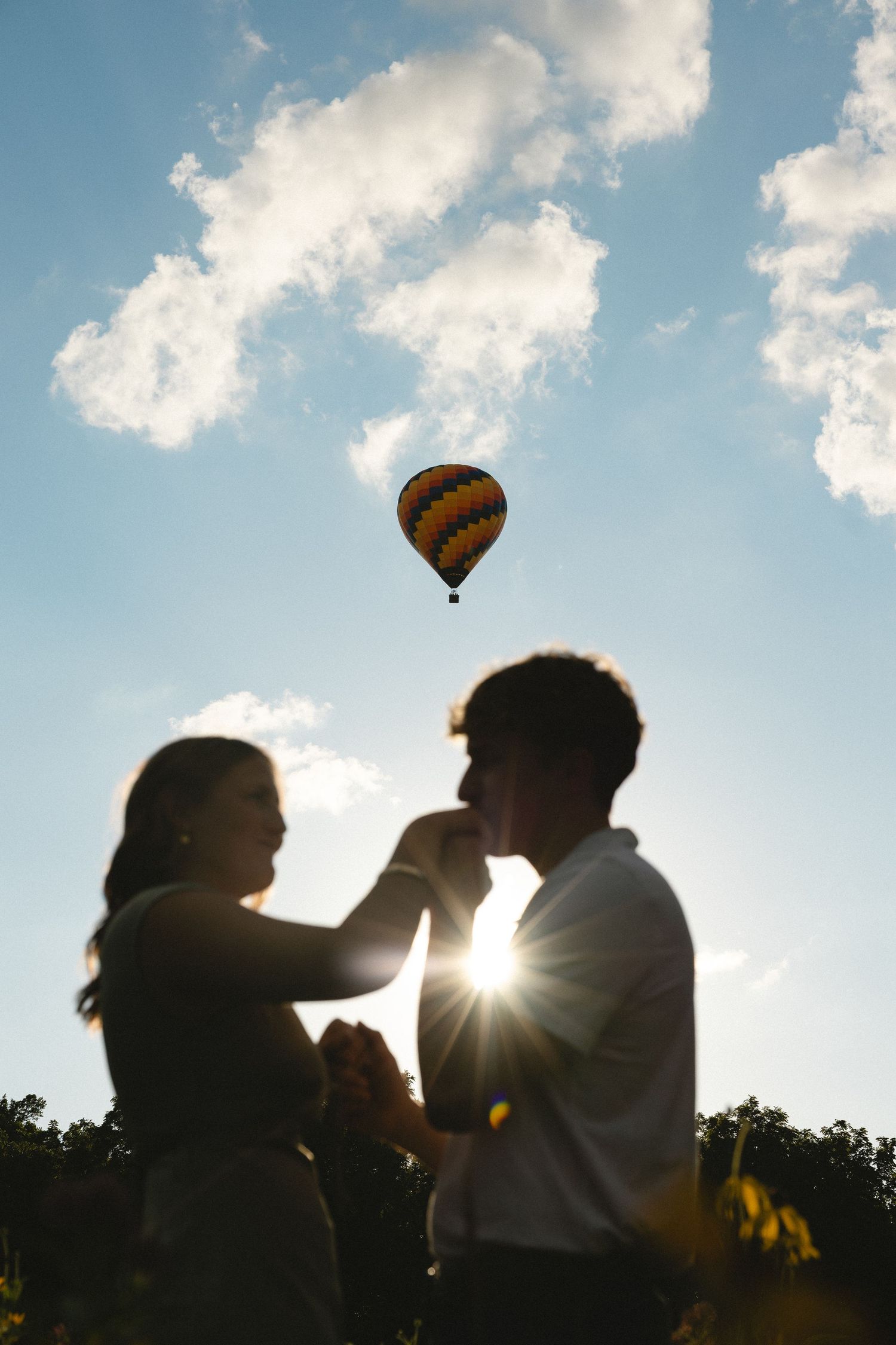 Romantic couple silhouetted against sunset sky with hot air balloon floating above, holding hands in a tender moment.