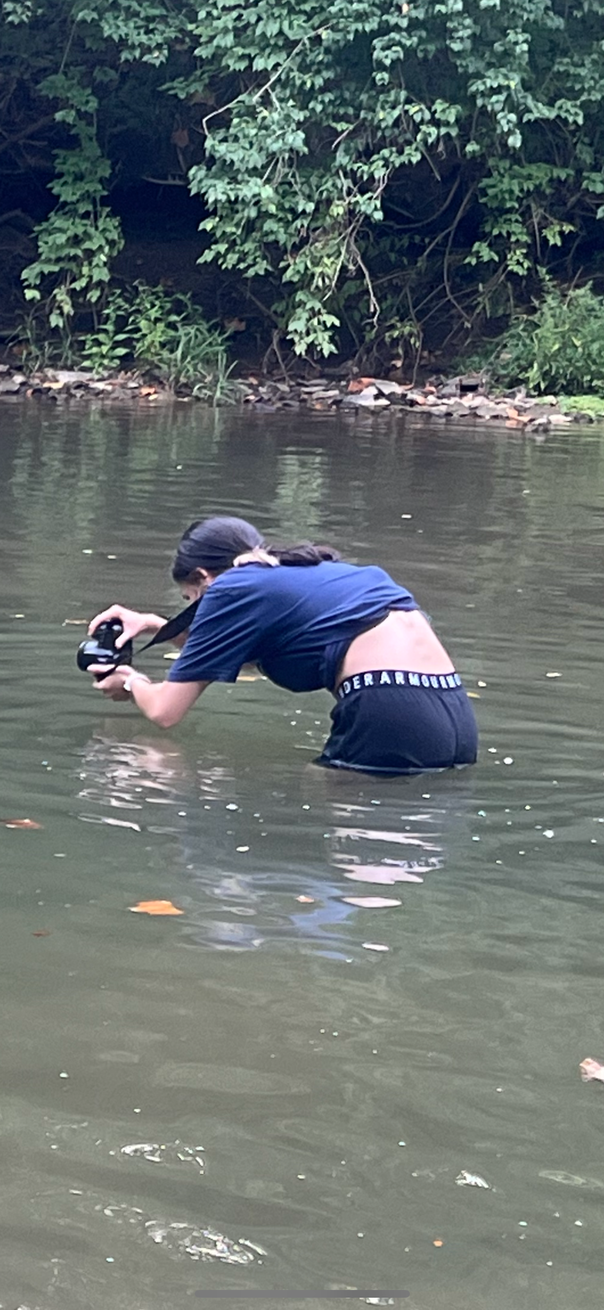 Person wading through murky river water wearing blue clothing holding a camera.