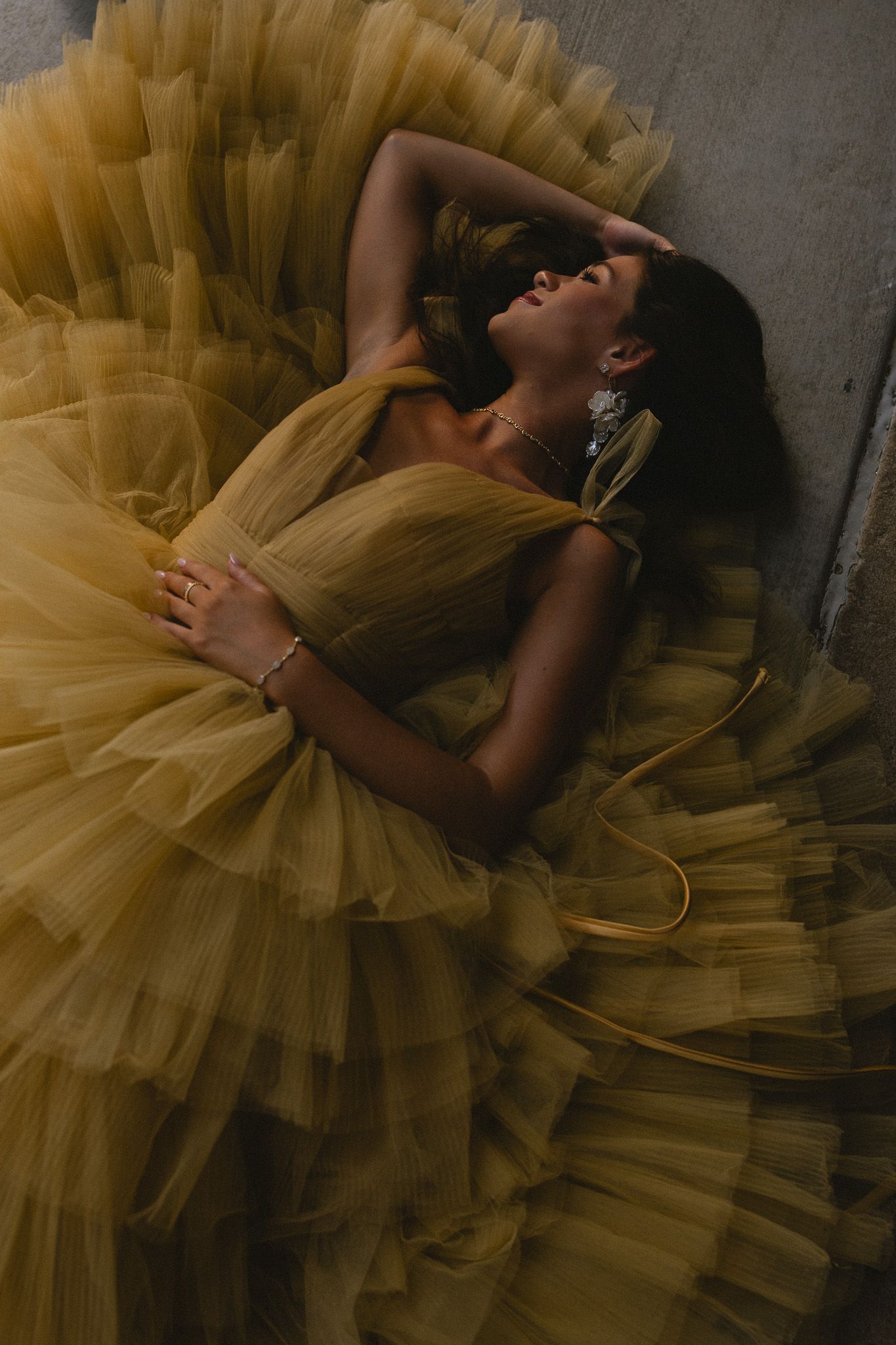Woman lying down in a voluminous golden yellow tulle ball gown, adorned with crystal earrings and bracelet.