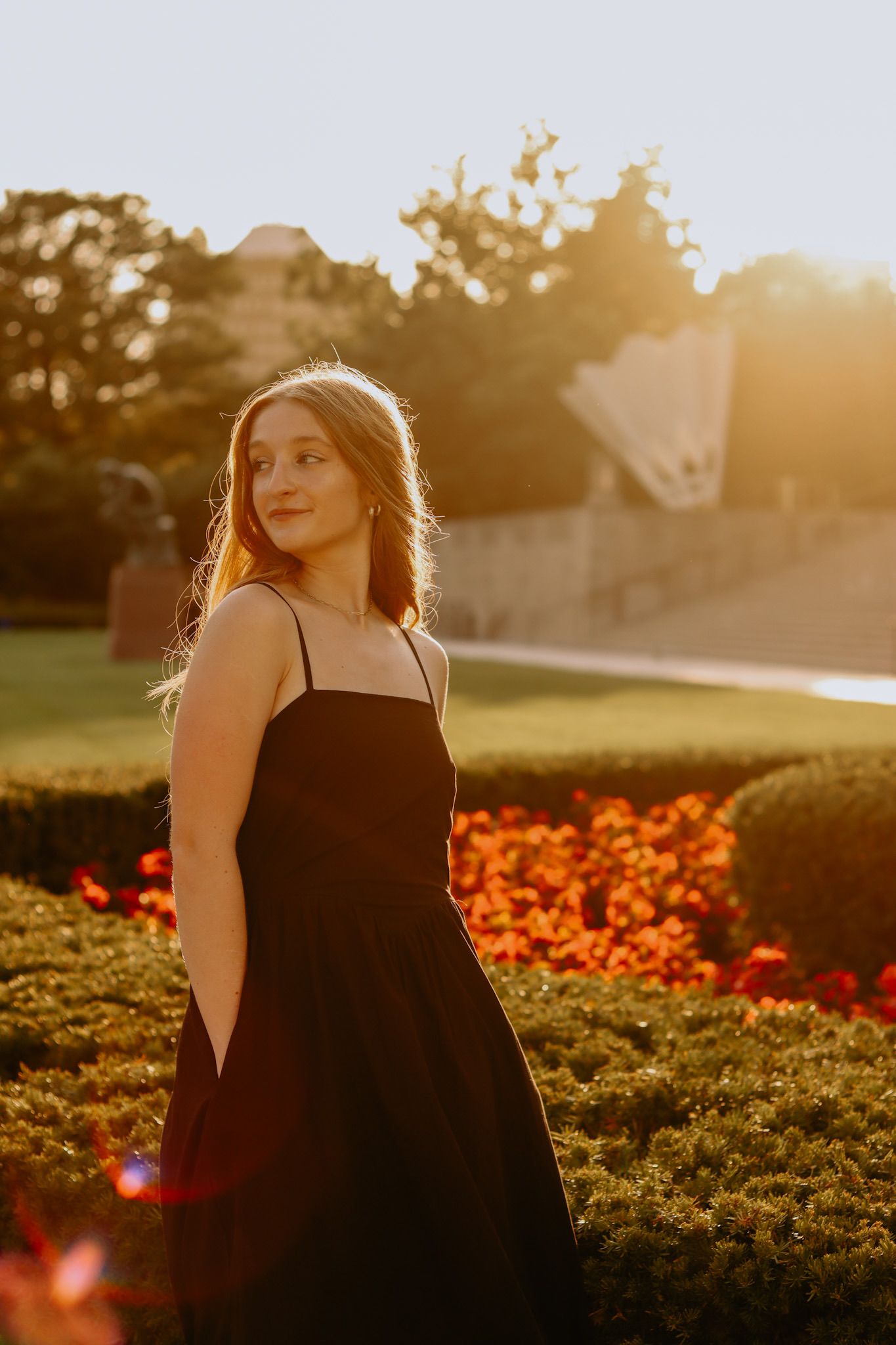 A golden hour portrait in a black dress near orange flowers and manicured bushes.