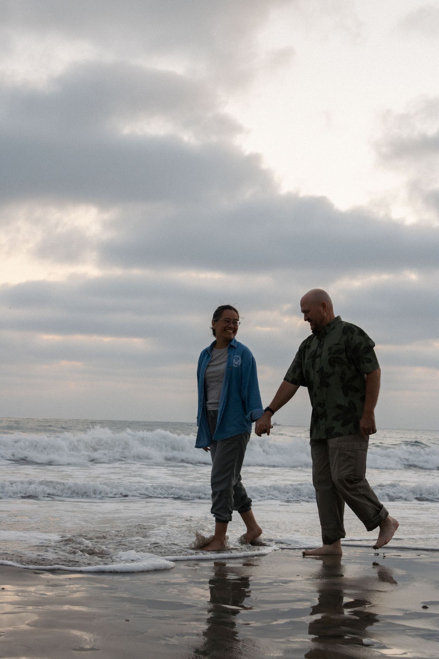 Two people wade together in ocean waves at sunset with cloudy skies.