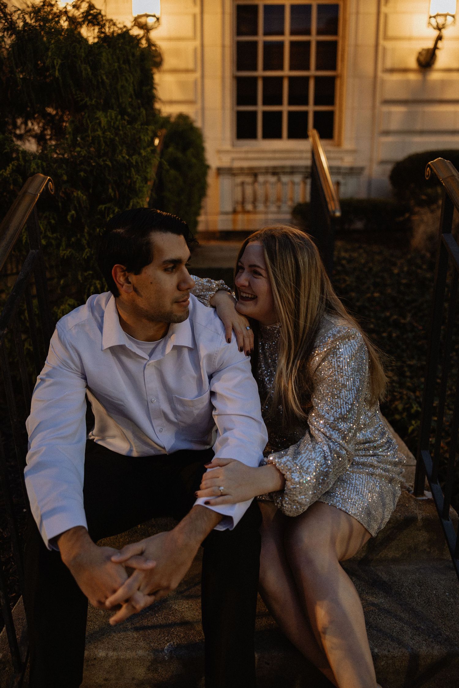 A romantic couple sits together at dusk on stone steps outside a building with warm lighting in the background.