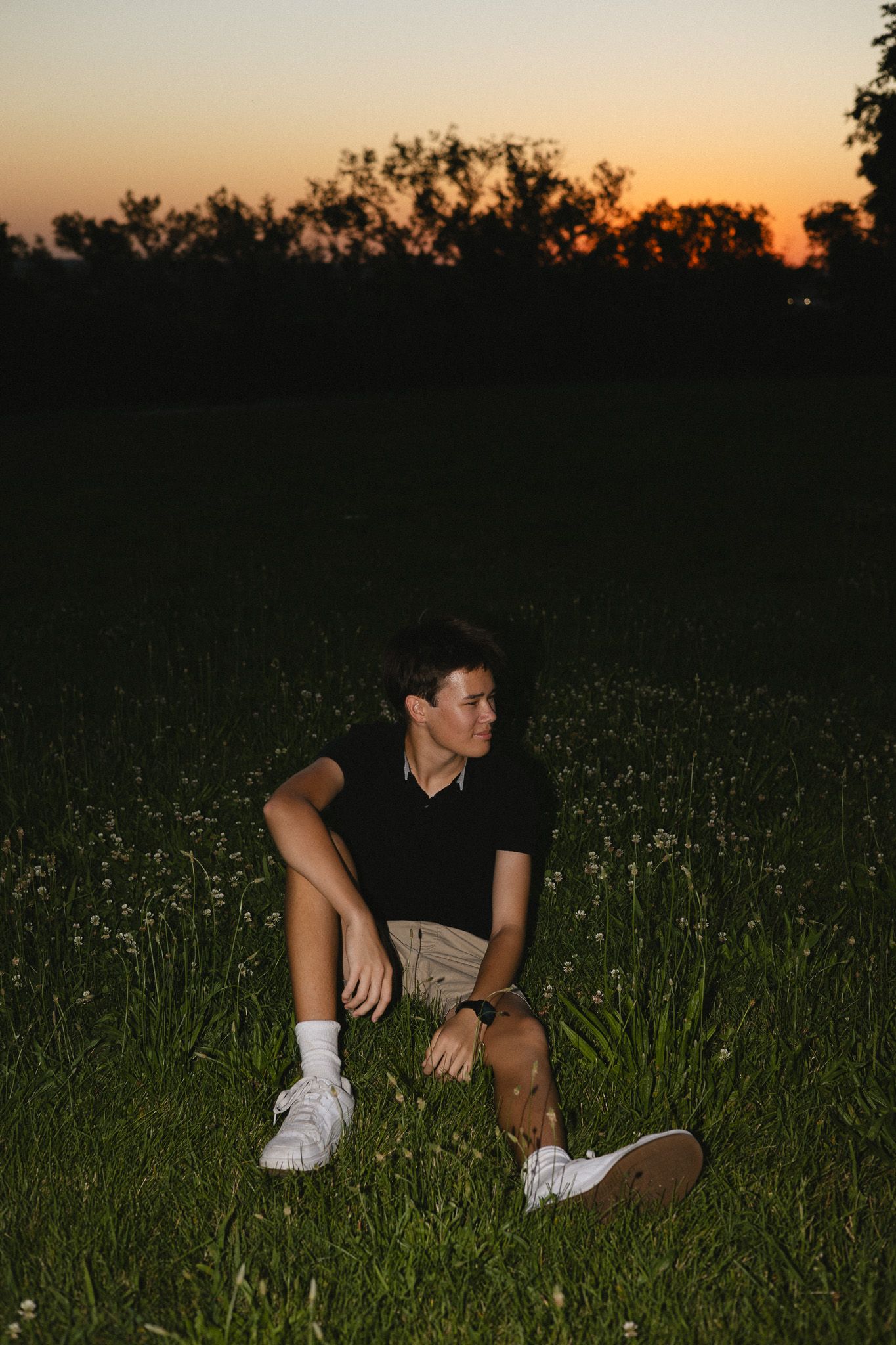 Young man sitting in a flower field at sunset, wearing black shirt and khaki shorts, smiling.