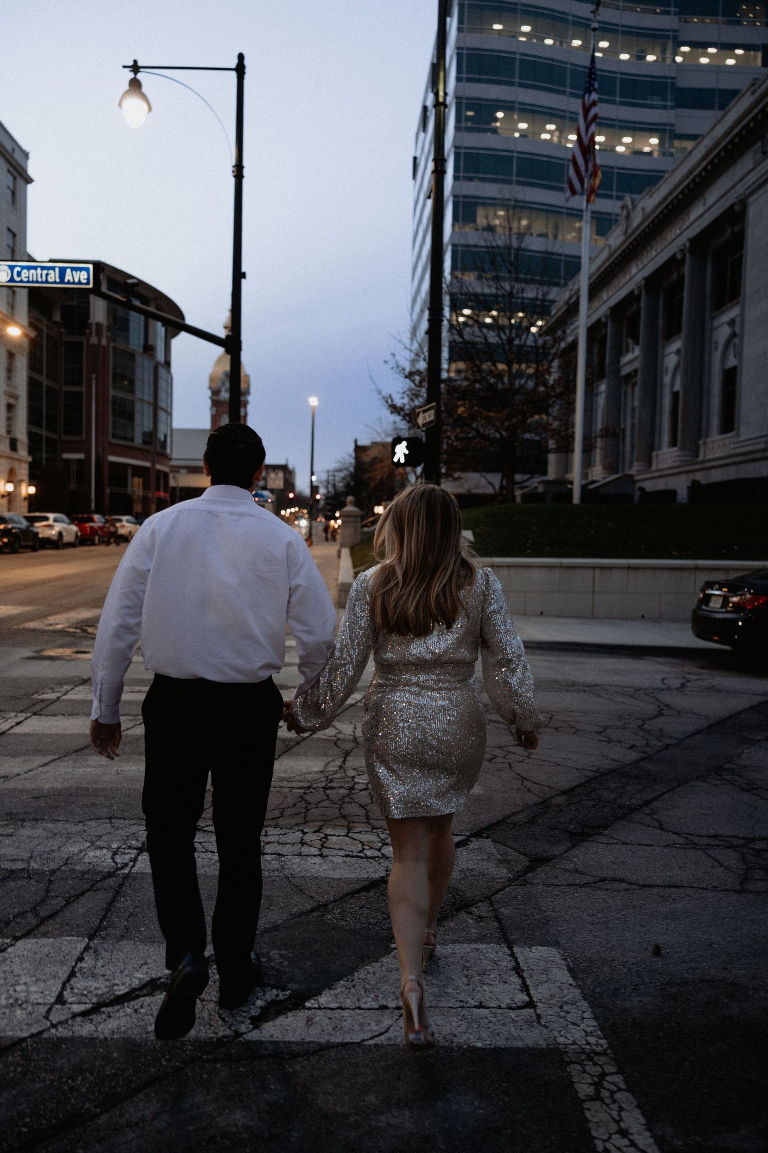A couple walking together at dusk on an urban street with tall buildings and street lights in the background.