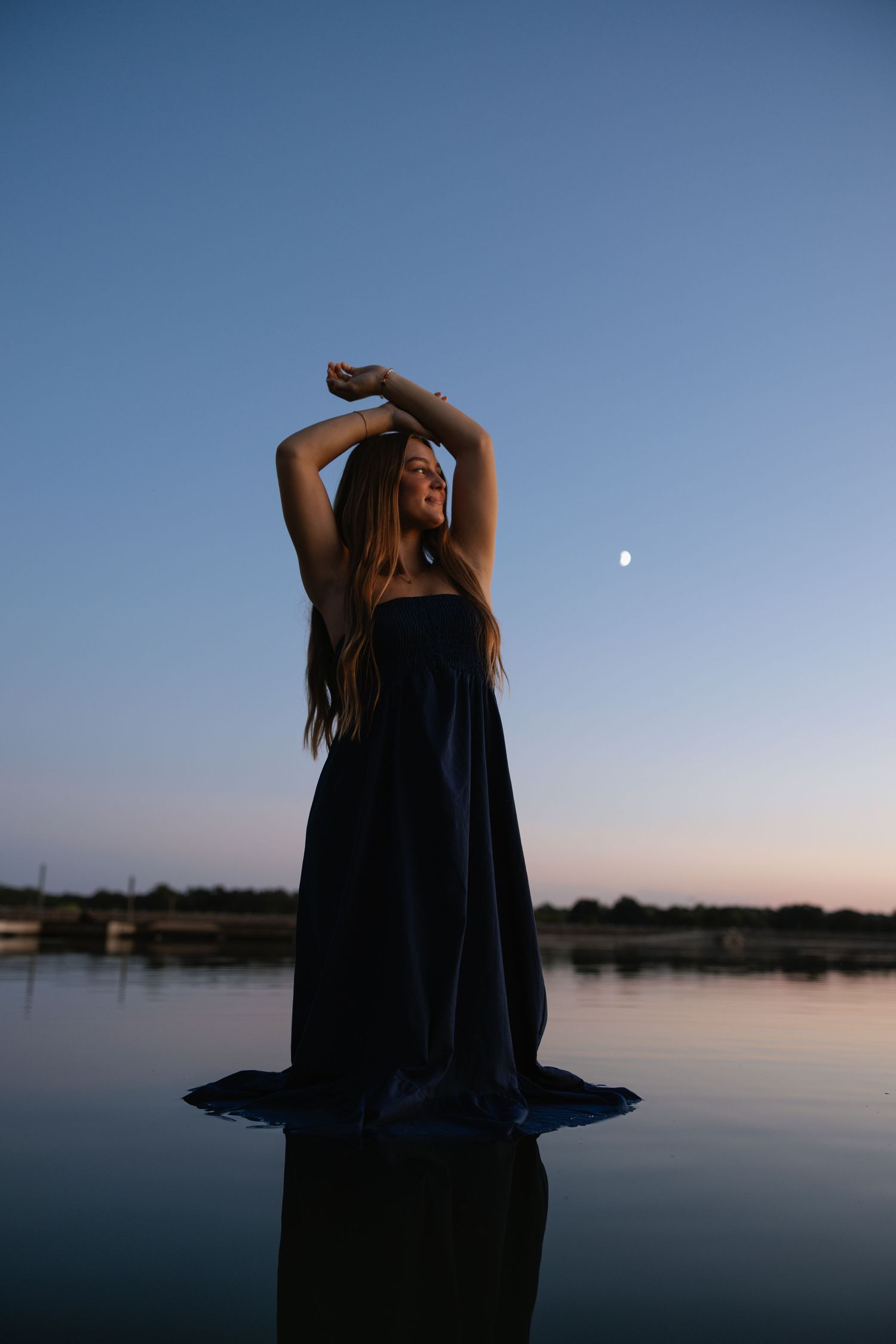 A silhouetted figure in a dress poses against a twilight sky with moon over water.