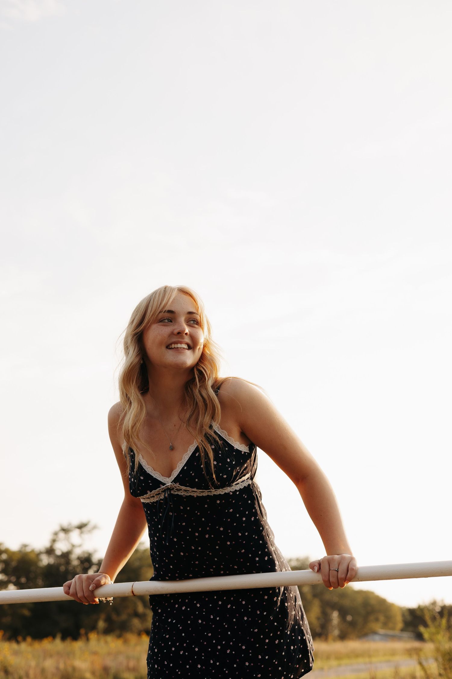 A person in a polka dot dress leans against a railing at sunset, laughing joyfully in a rural setting.
