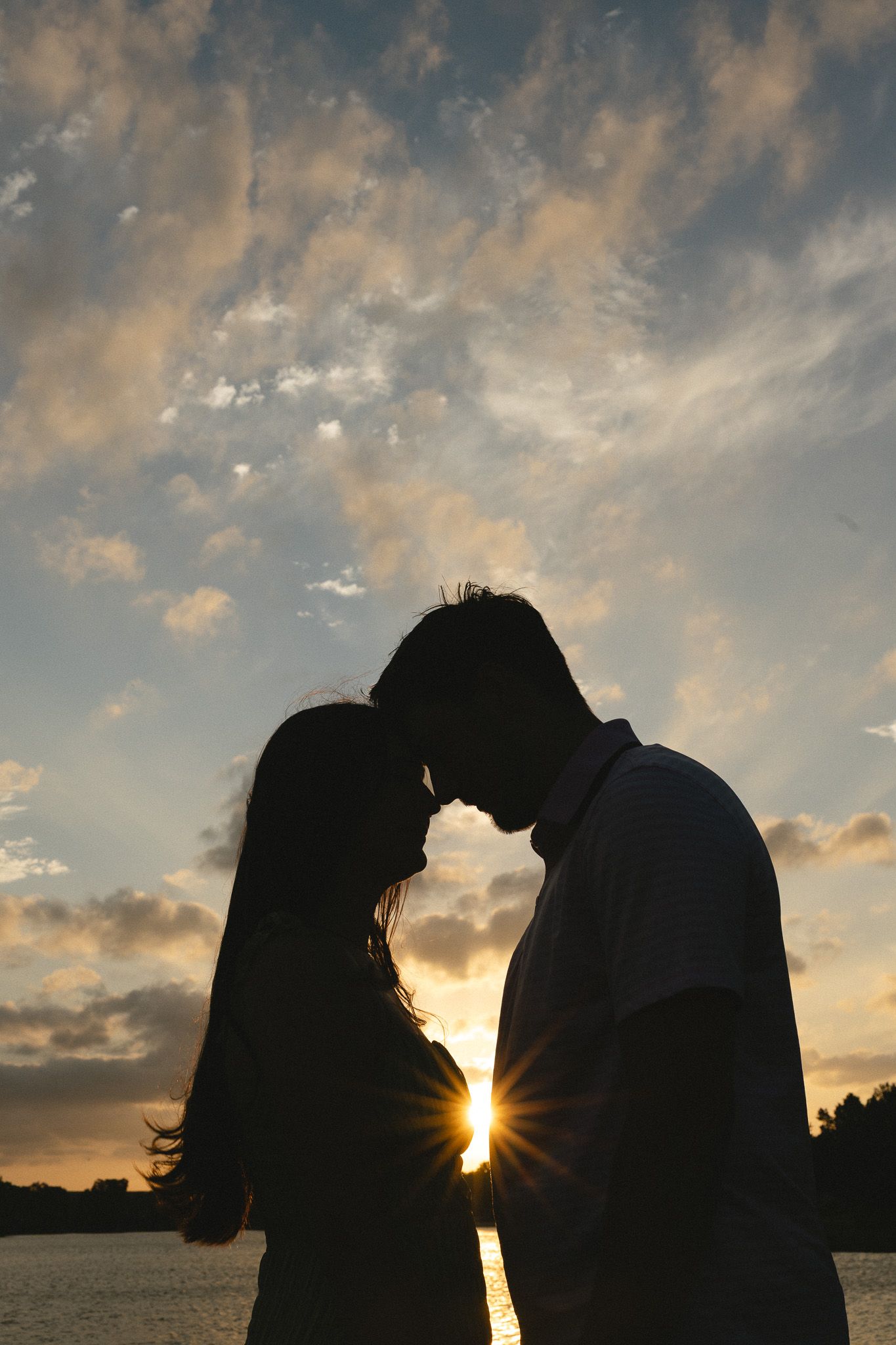 Romantic silhouette of a couple touching foreheads at sunset with dramatic cloudy sky and sunburst behind them.
