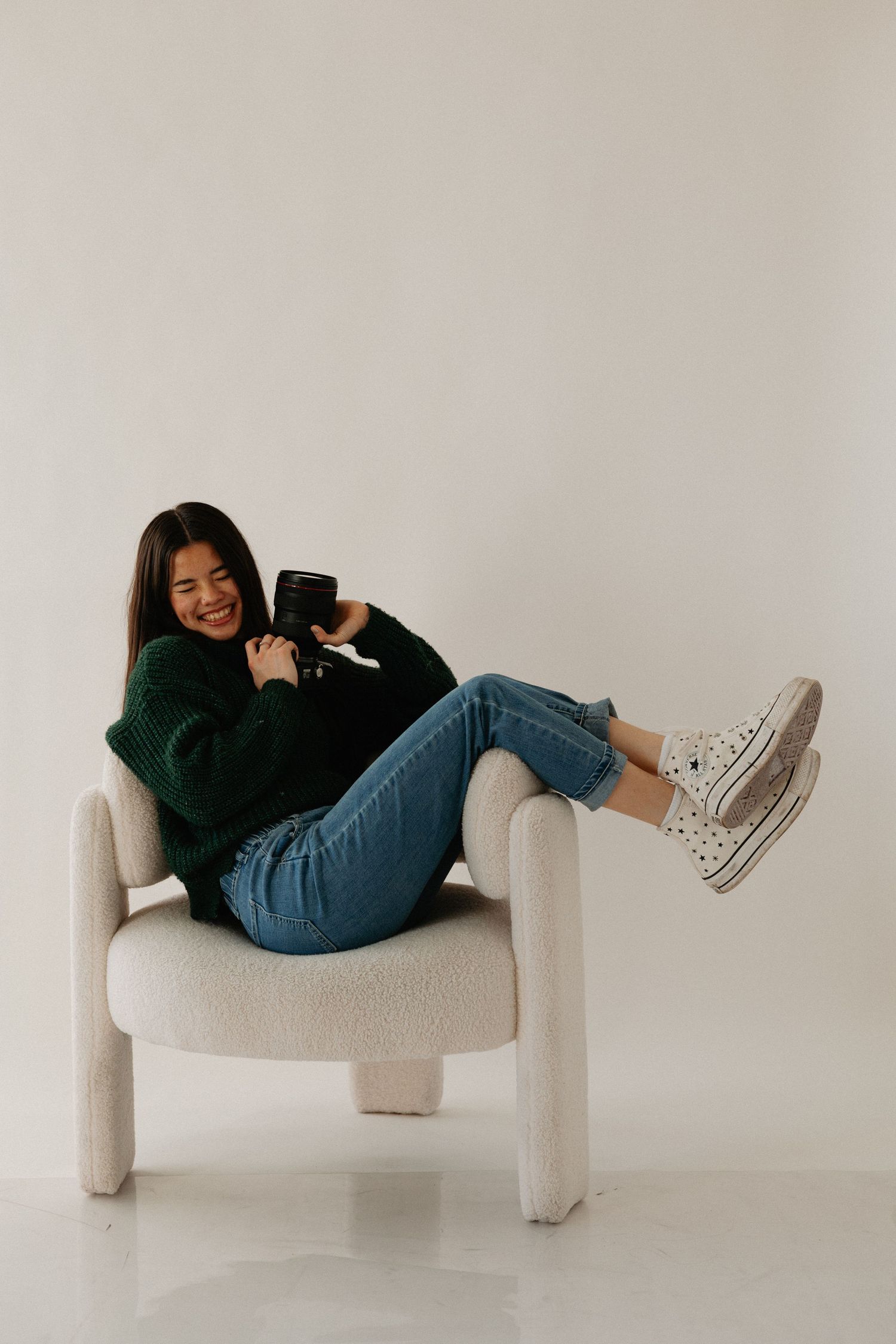 Playful studio portrait on modern white chair wearing black sweater and jeans.