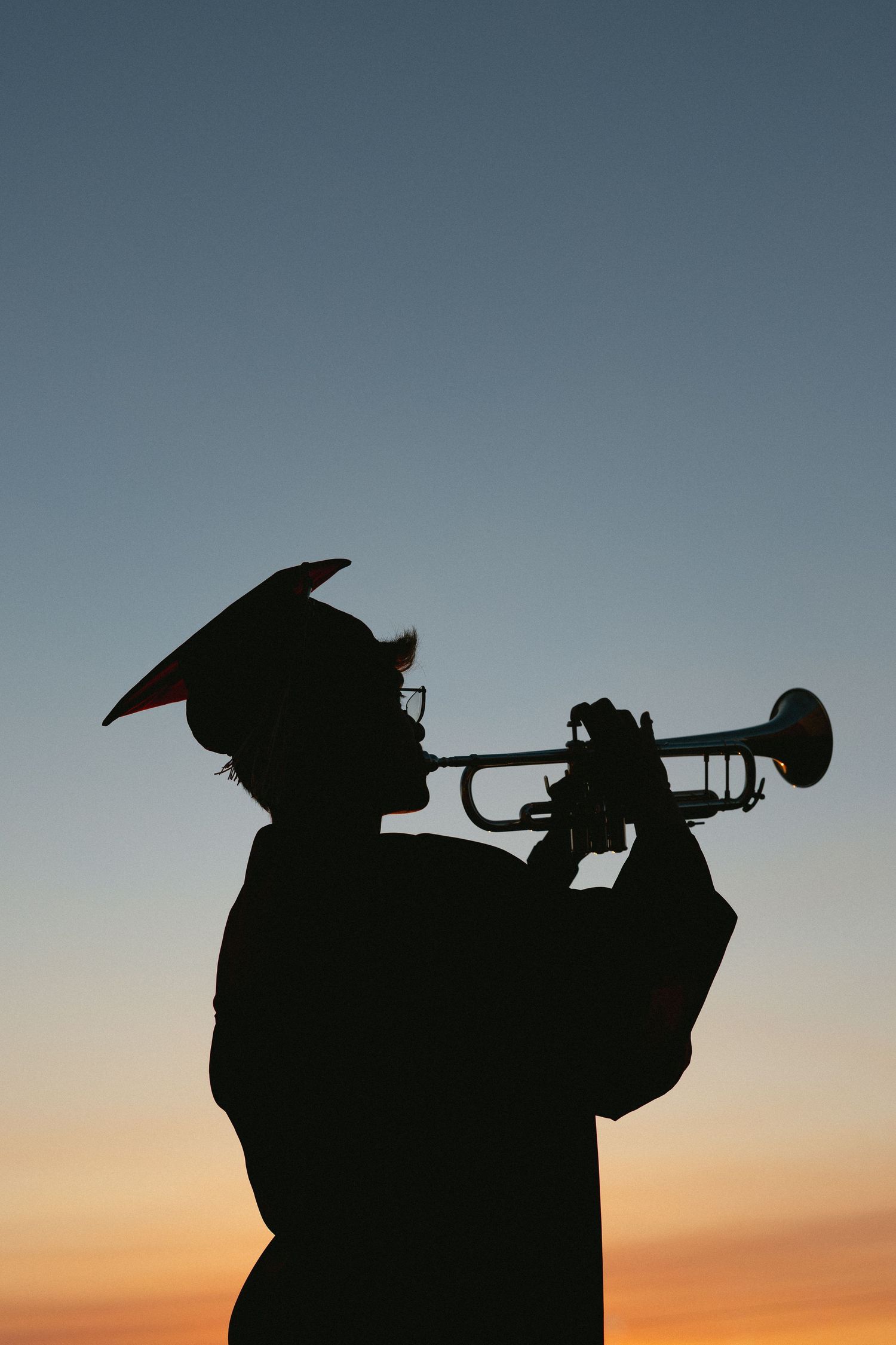Silhouette of a graduate in cap and gown playing trumpet against a colorful sunset sky.