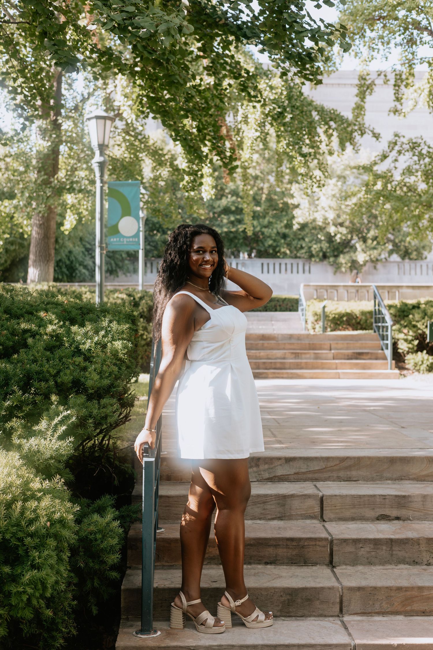 Young Black woman in white mini dress and heels posing on outdoor steps surrounded by green trees.