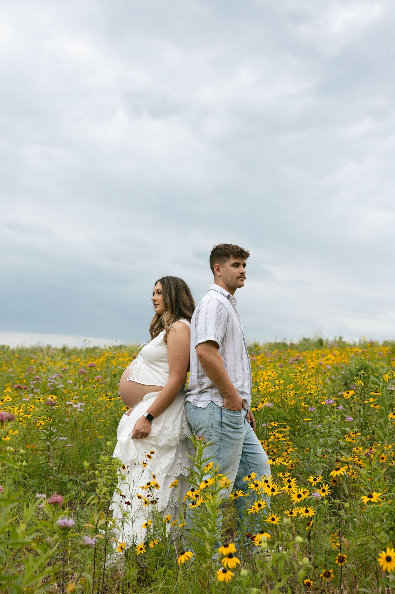 Pregnant couple standing back-to-back in a wildflower field with yellow blooms under a cloudy sky.