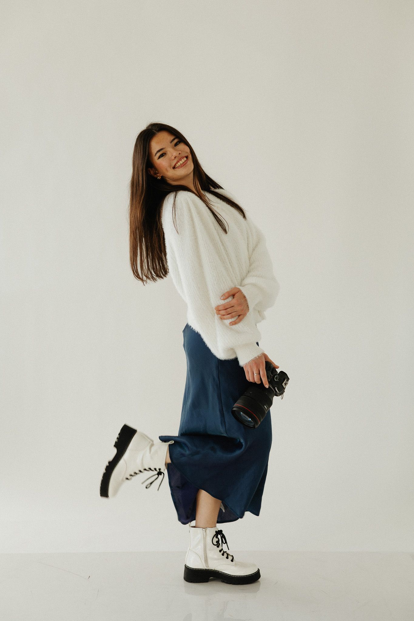 Model in white sweater and jeans poses playfully against white background in fashion photoshoot series.