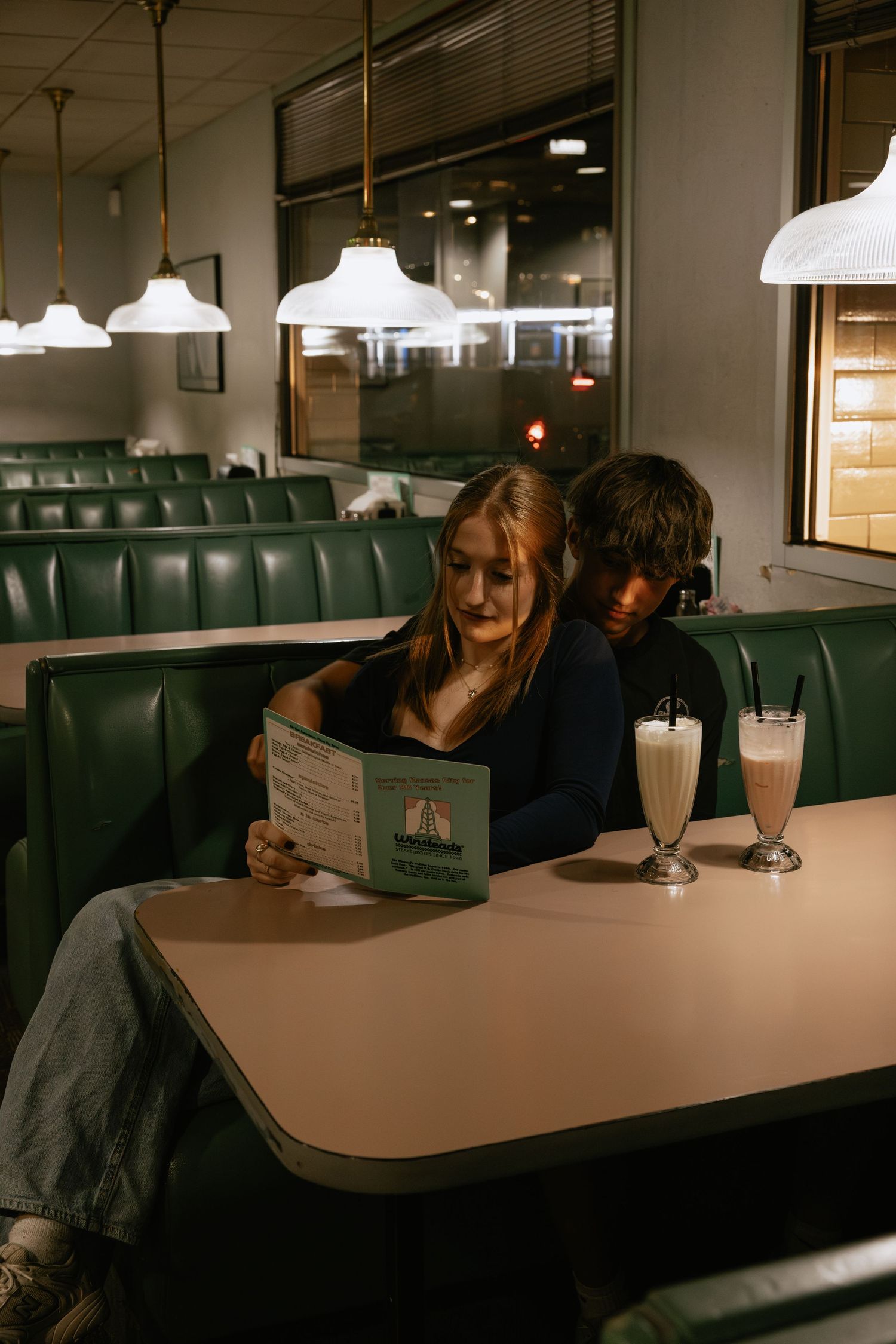 Couple sharing milkshakes at retro diner booth with pendant lighting.