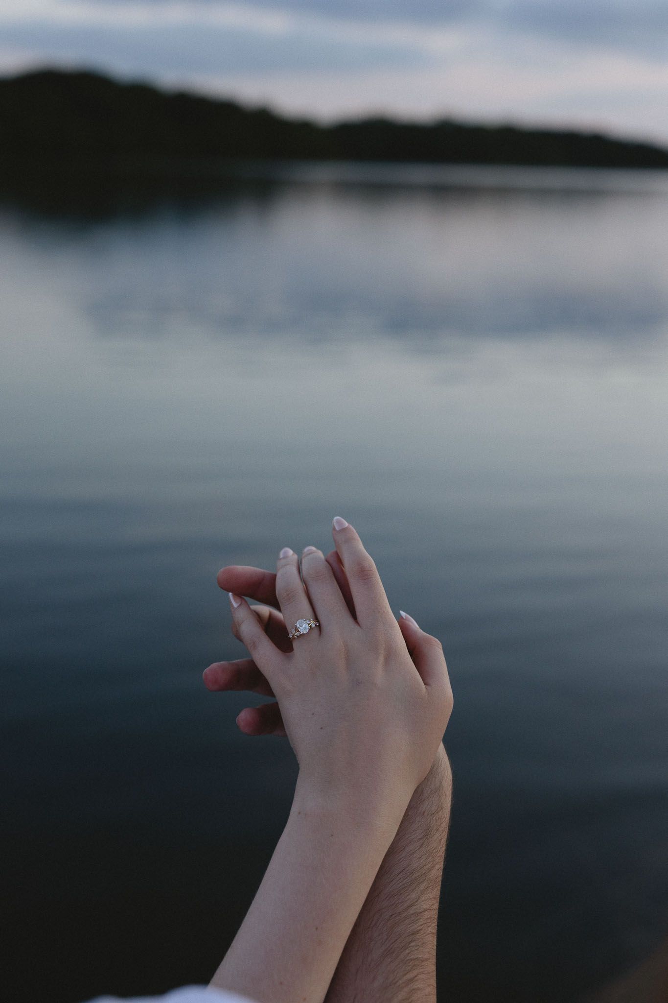 Two hands touching near a lake at dusk, one wearing a diamond engagement ring, symbolizing a romantic proposal.