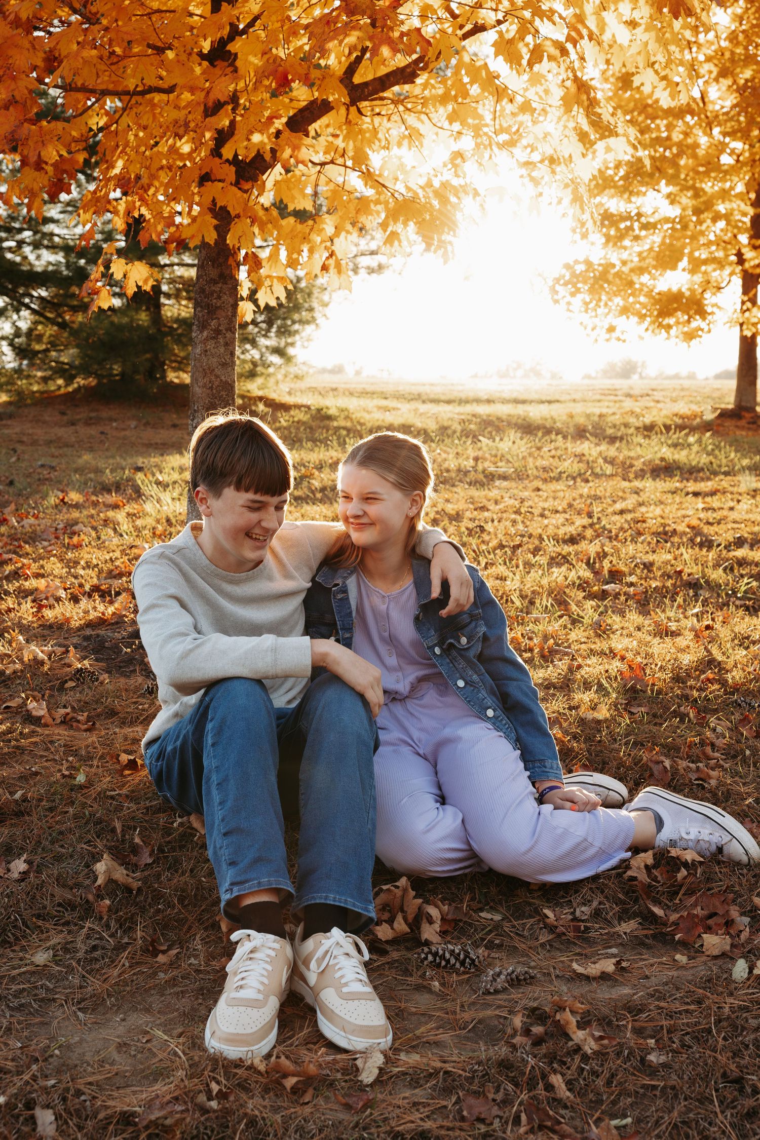 Two siblings sits together on fallen autumn leaves during golden hour sunset, sharing a tender moment of laughter.
