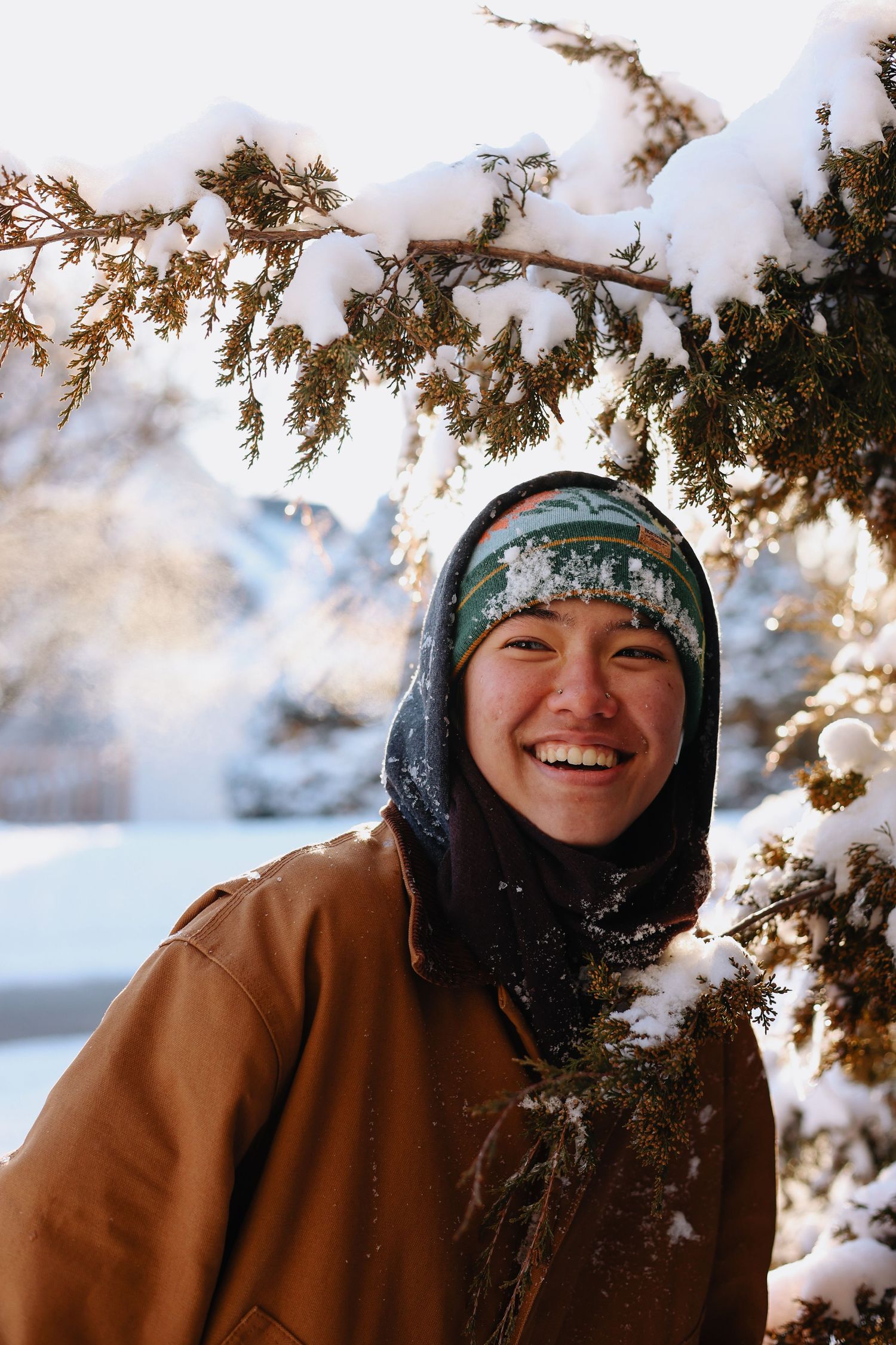 Person wearing winter hat and brown coat smiles while surrounded by snowy evergreen branches in winter scene.