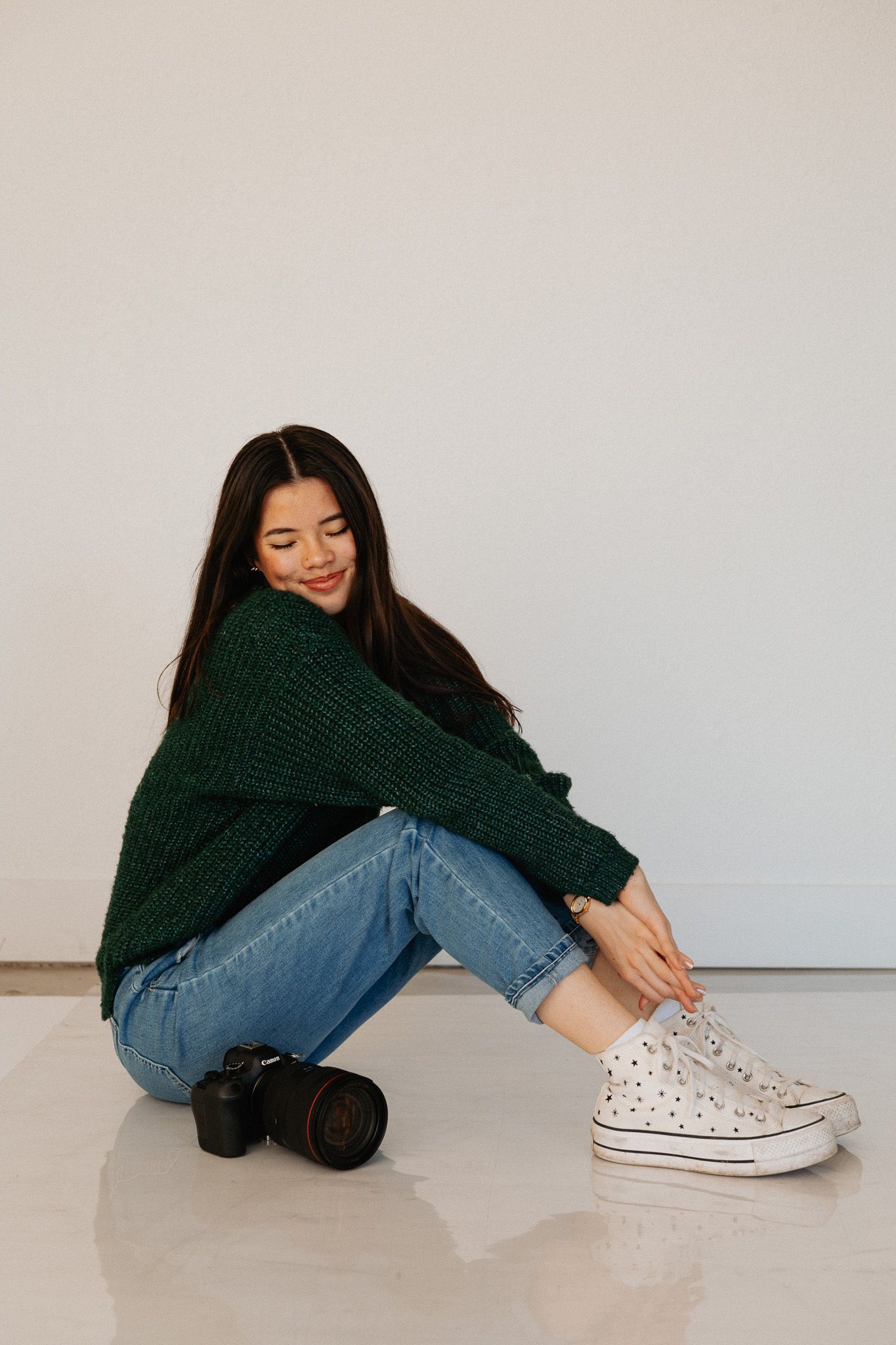 Person in green sweater and jeans sits casually on floor wearing white patterned sneakers.