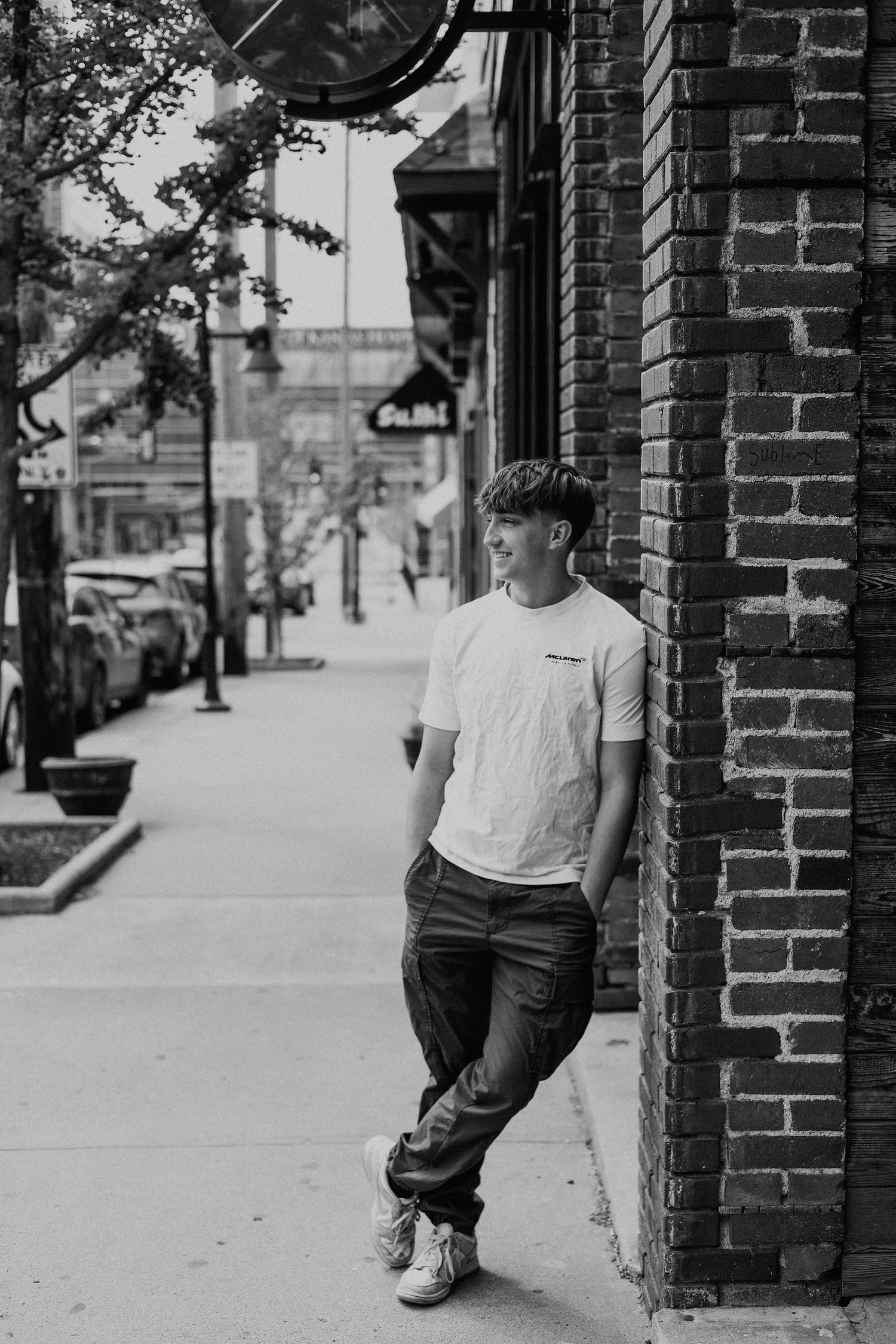 Black and white street photo of someone leaning against a brick building wall wearing a white t-shirt and jeans.