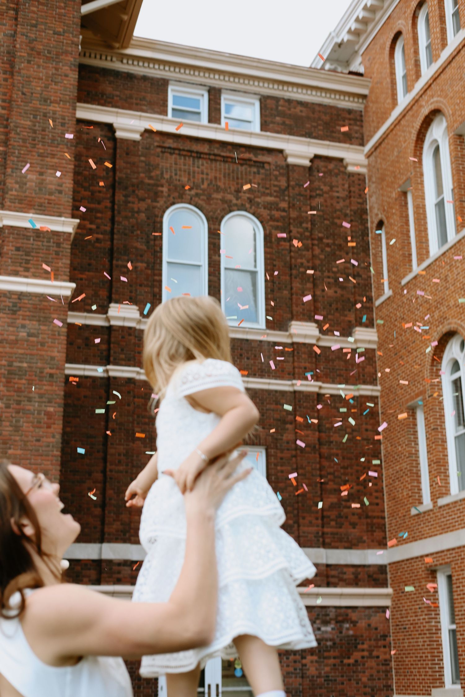 People in white dresses celebrating with confetti in front of brick building.