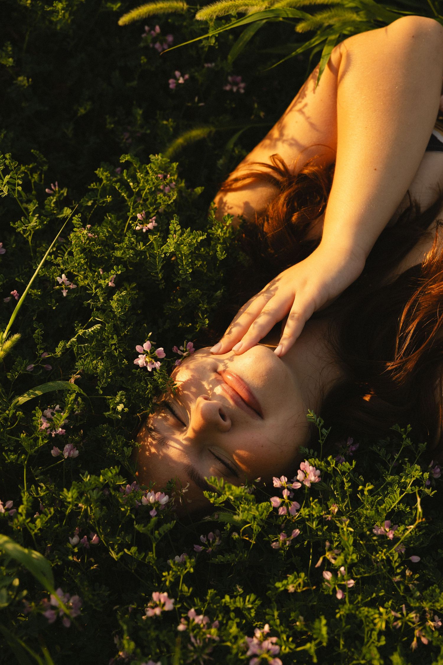 A young woman with red hair lies peacefully in a field of wildflowers, hand resting on her face in golden hour light.