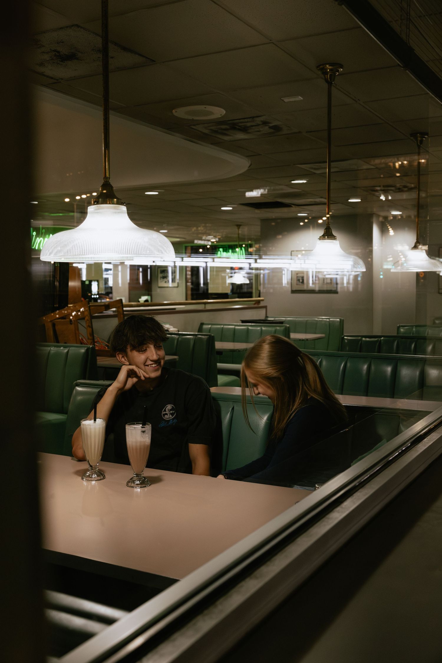 Two people share milkshakes in a retro diner with vintage pendant lights and turquoise booths.