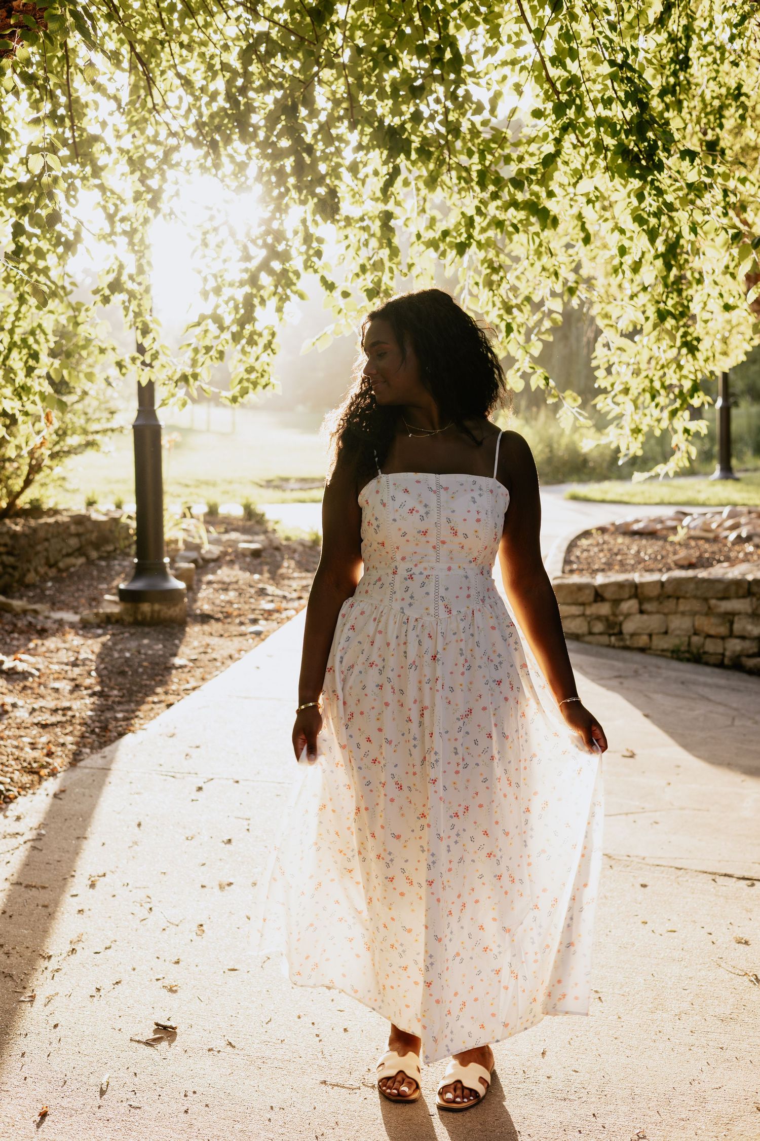 Girl looking off to side while holding dress, standing on walkway with sun shining through leaves behind her.