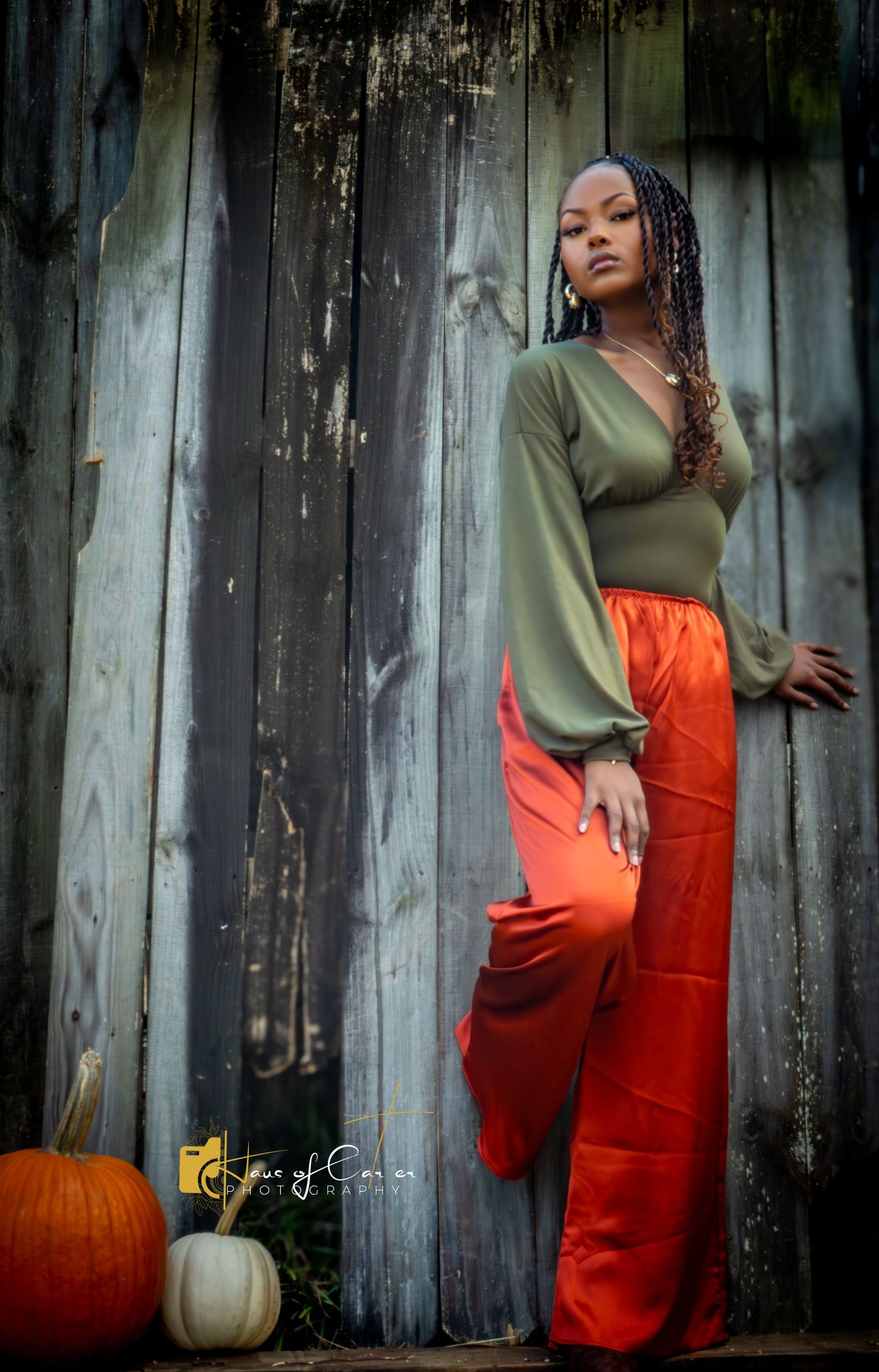 Fall fashion portrait with orange pants and olive top against weathered wooden fence with pumpkin decor.