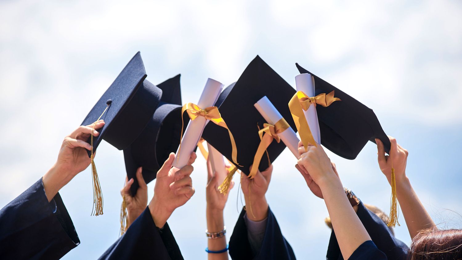 Graduates in black caps and gowns raise their diplomas in celebration against a bright blue sky.