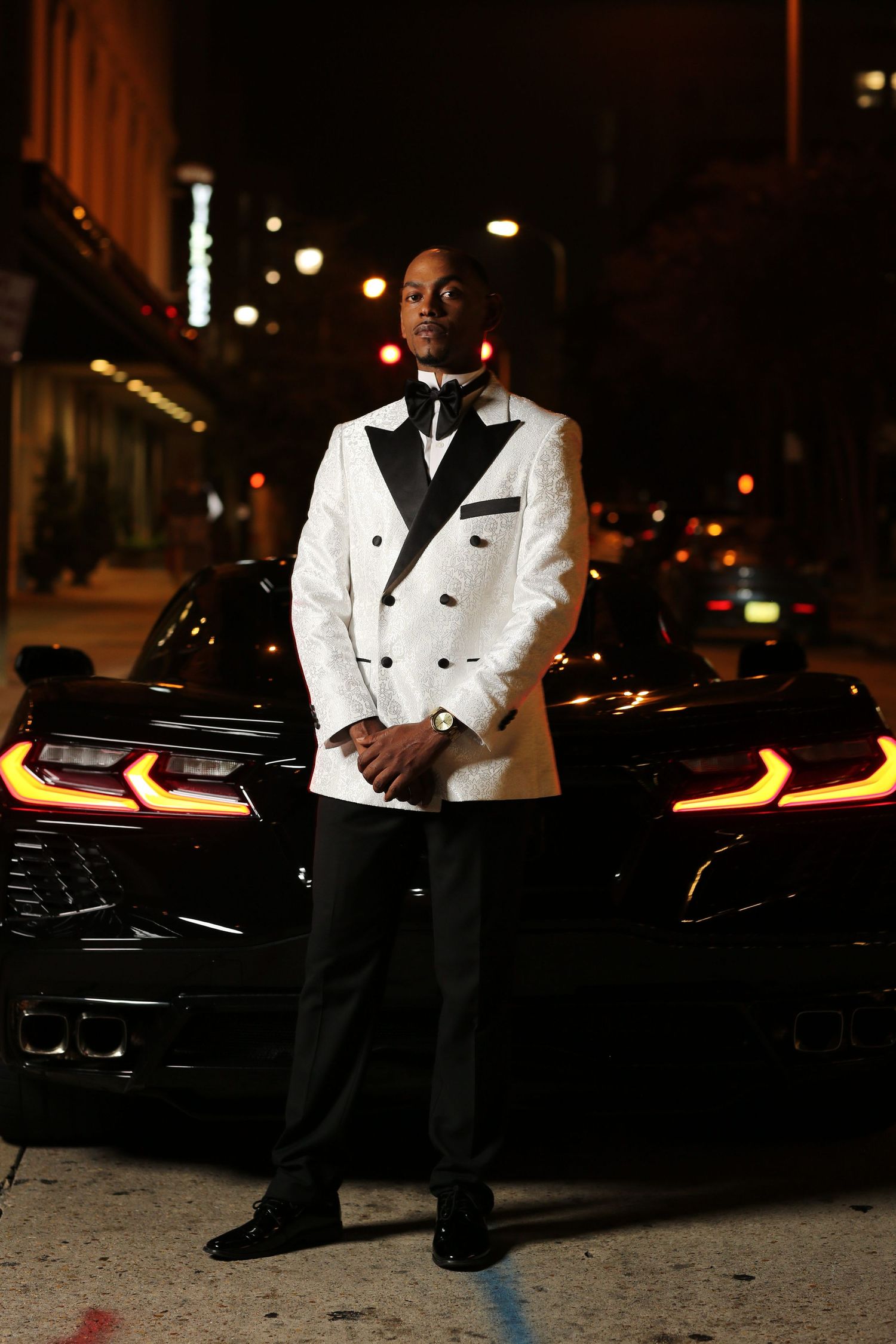 A person in a white tuxedo jacket and black pants poses elegantly against a dark car at night.