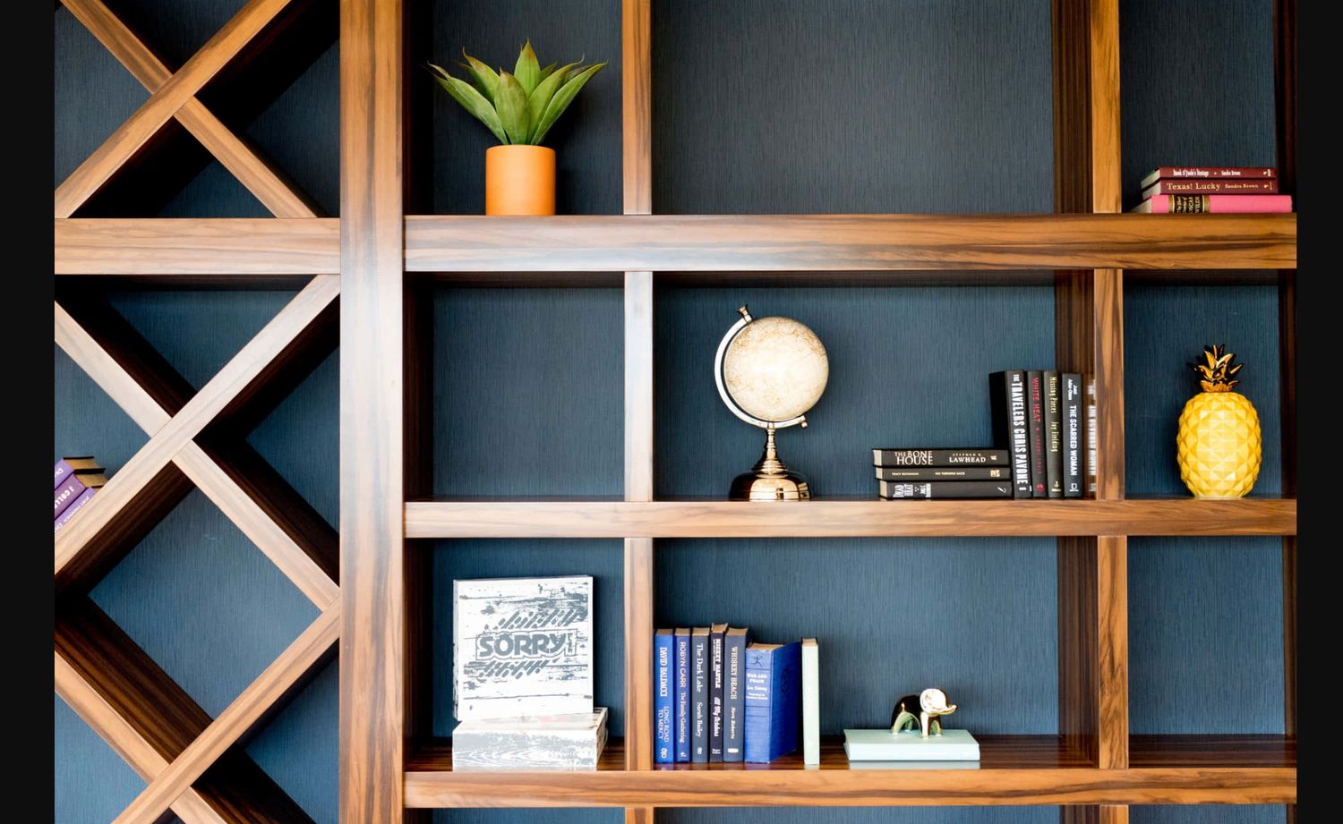 Modern wooden shelving unit with diagonal cross supports mounted on dark blue wall displaying books and decorative items.