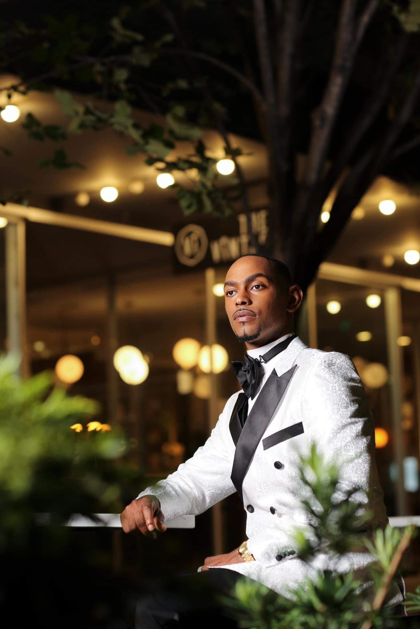 A person in a white tuxedo jacket and black bow tie sits at an elegant evening event with string lights overhead.