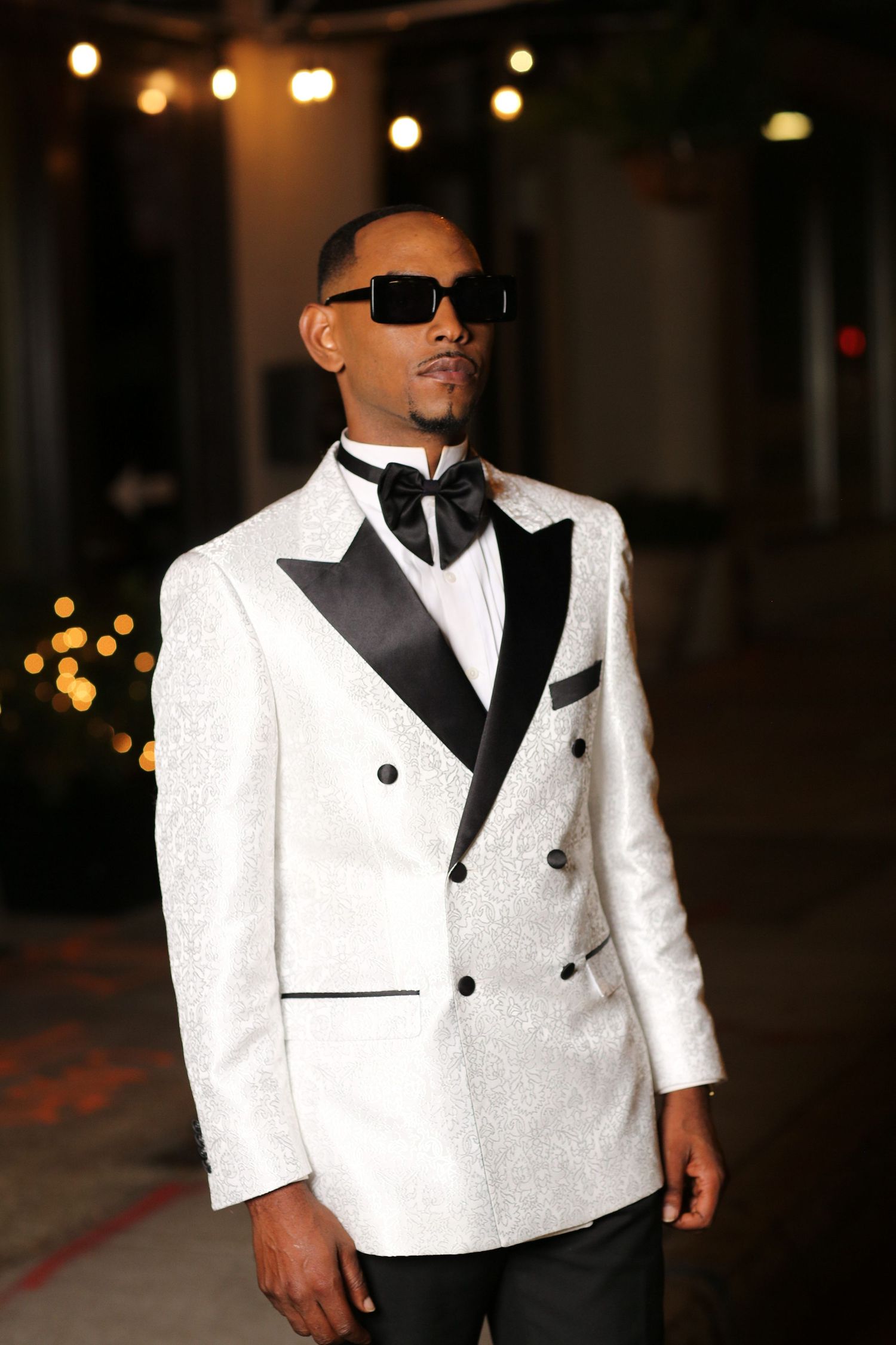 Stylish double-breasted white tuxedo jacket with black lapels and bow tie worn with sunglasses in evening setting.
