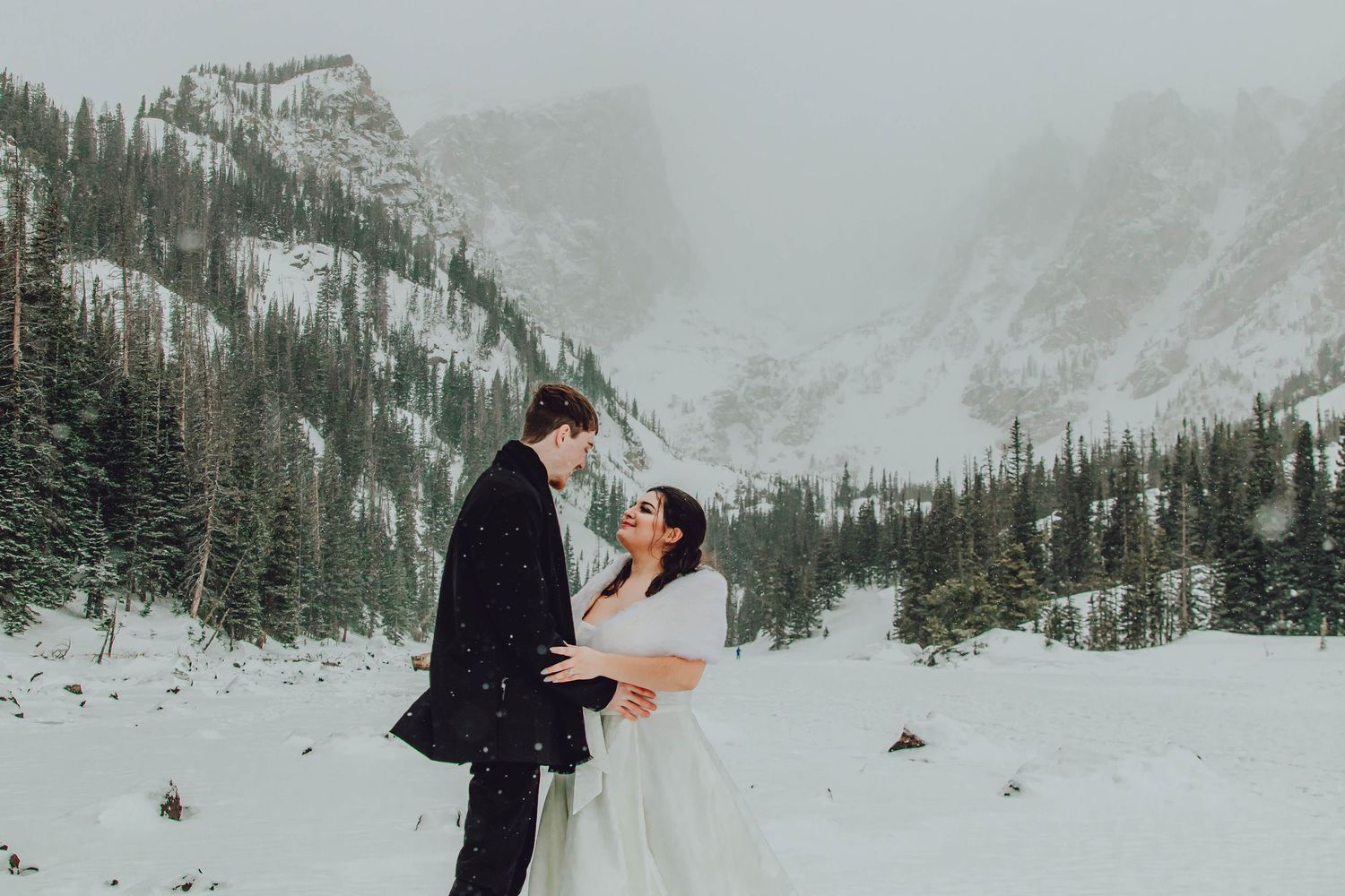 A couple embraces during their winter wedding ceremony in a snowy mountain valley surrounded by evergreen trees.