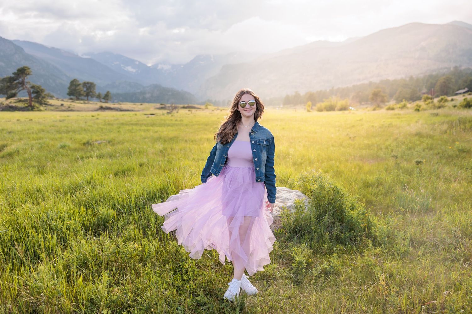 A person in a pink tulle skirt and denim jacket twirls in a sunlit mountain meadow at golden hour.