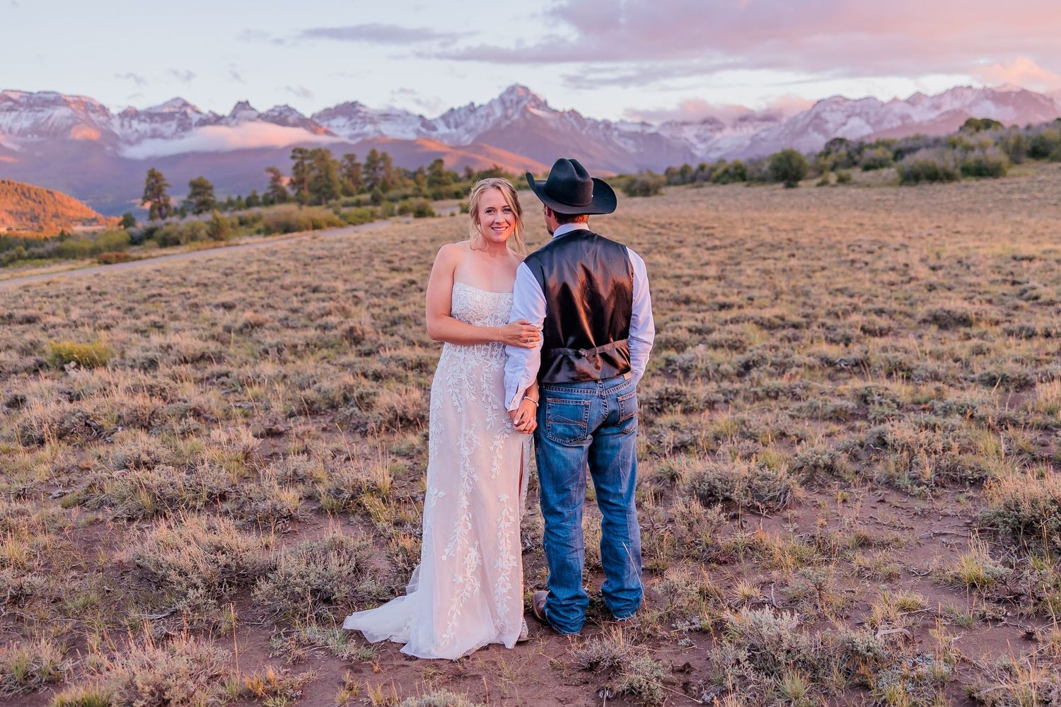 A romantic couple poses against a desert landscape with mountains and pink sunset sky in the background.