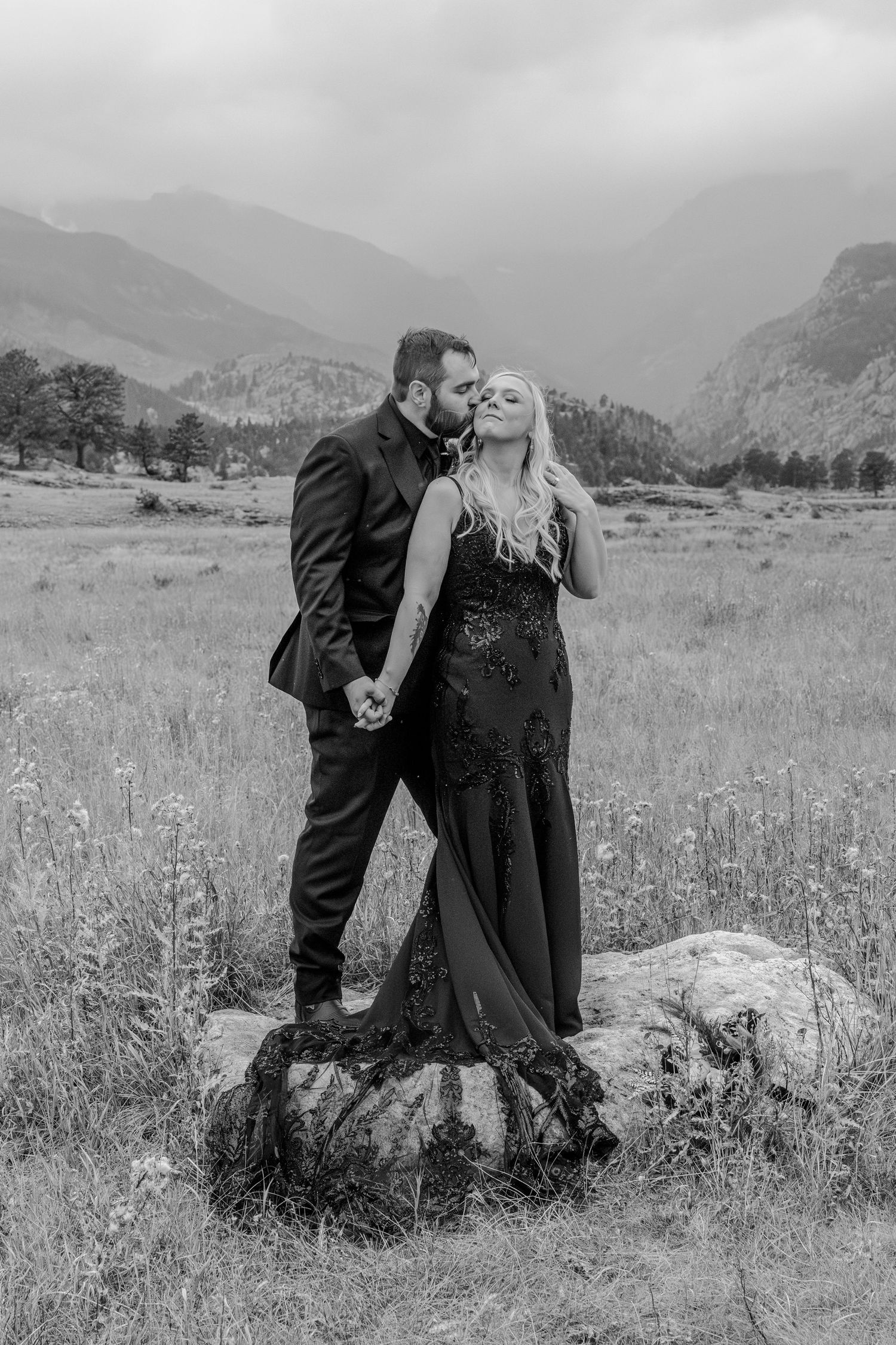 A romantic couple in formal attire embraces in a mountain meadow field with wildflowers and scenic peaks in the background.