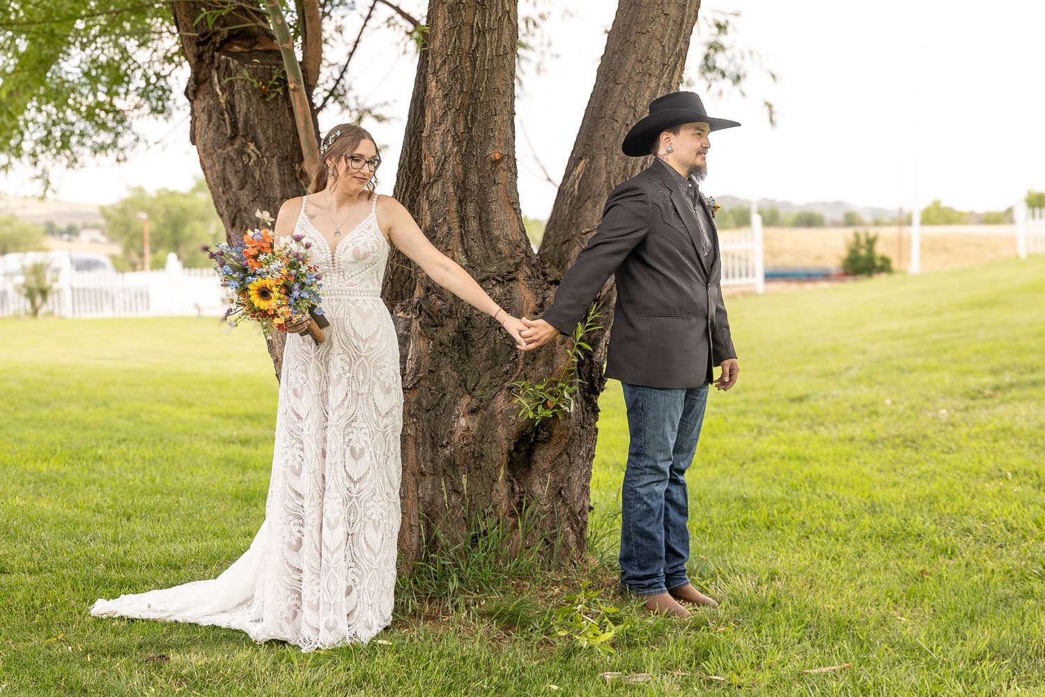 A romantic wedding photo of a couple posing playfully on opposite sides of a tree trunk in a grassy field.