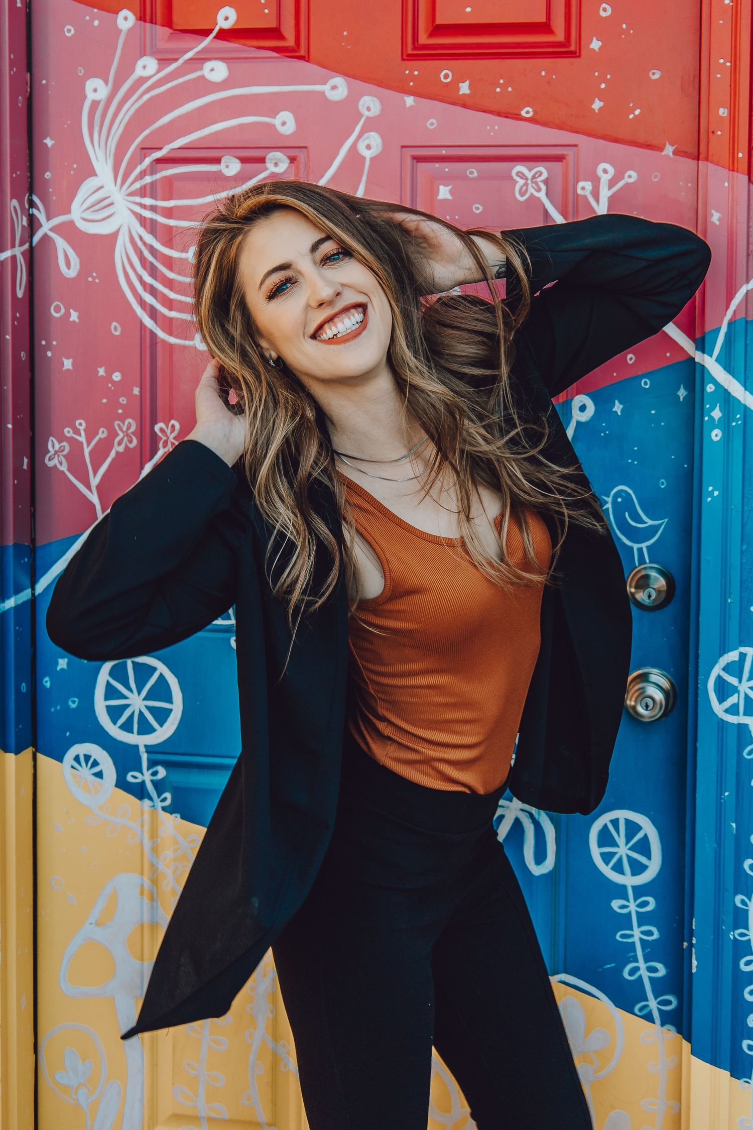 Woman in orange top and black jacket poses playfully against colorful wall with whimsical painted designs.