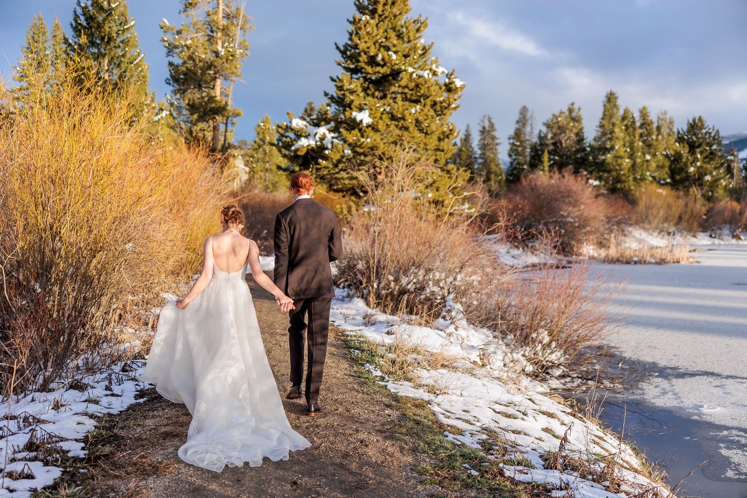 A winter wedding couple walks through a snowy landscape with evergreen trees and golden sunlight.