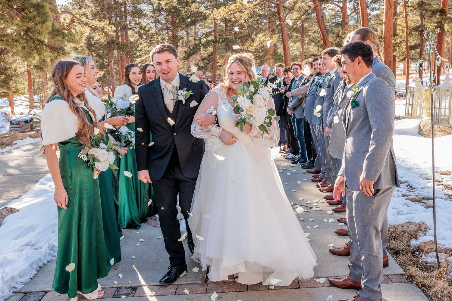 Winter wedding ceremony with wedding party in green dresses and gray suits among snow-covered pine trees.