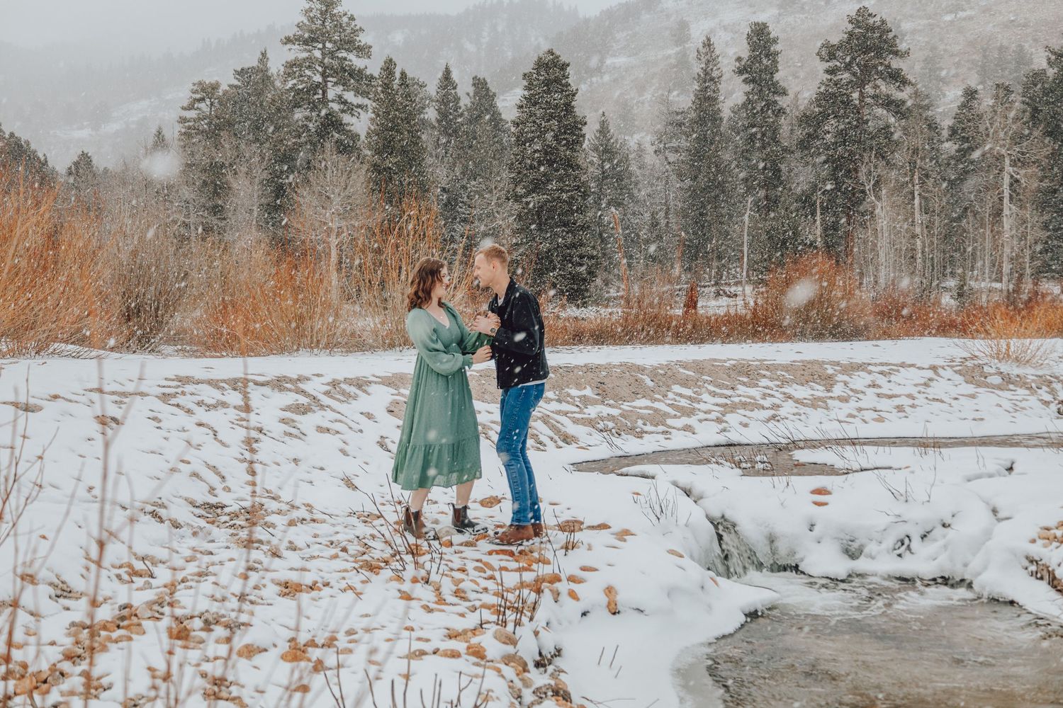 A winter engagement photo session in a snowy mountain landscape with evergreen trees and warm earth tones.
