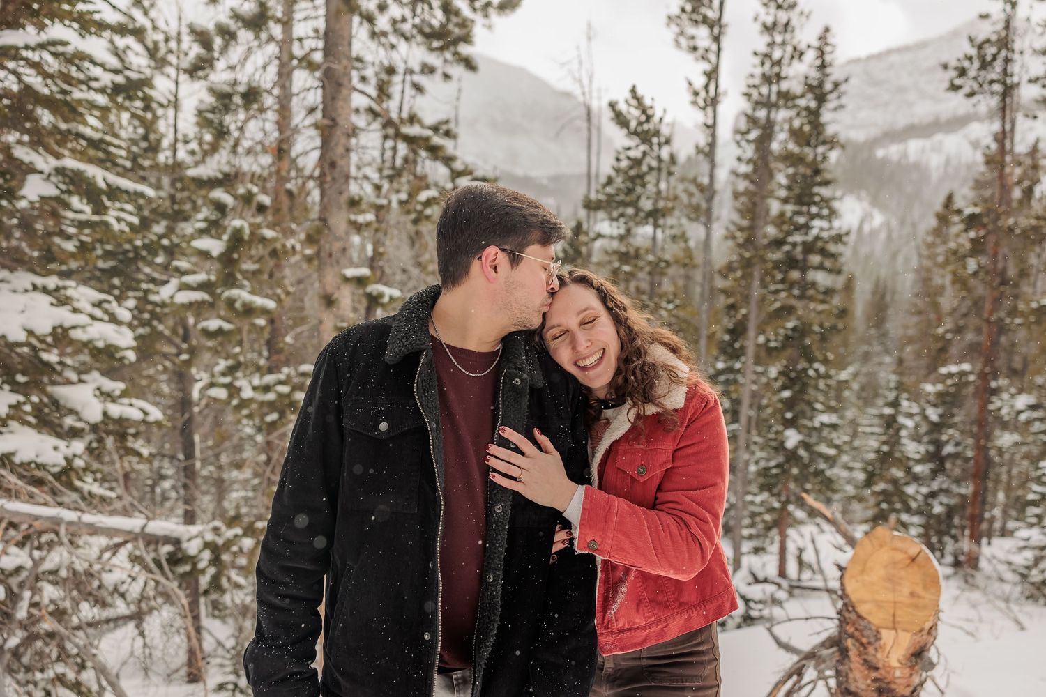 A couple shares romantic moments in a snowy forest setting during winter, surrounded by evergreen trees.