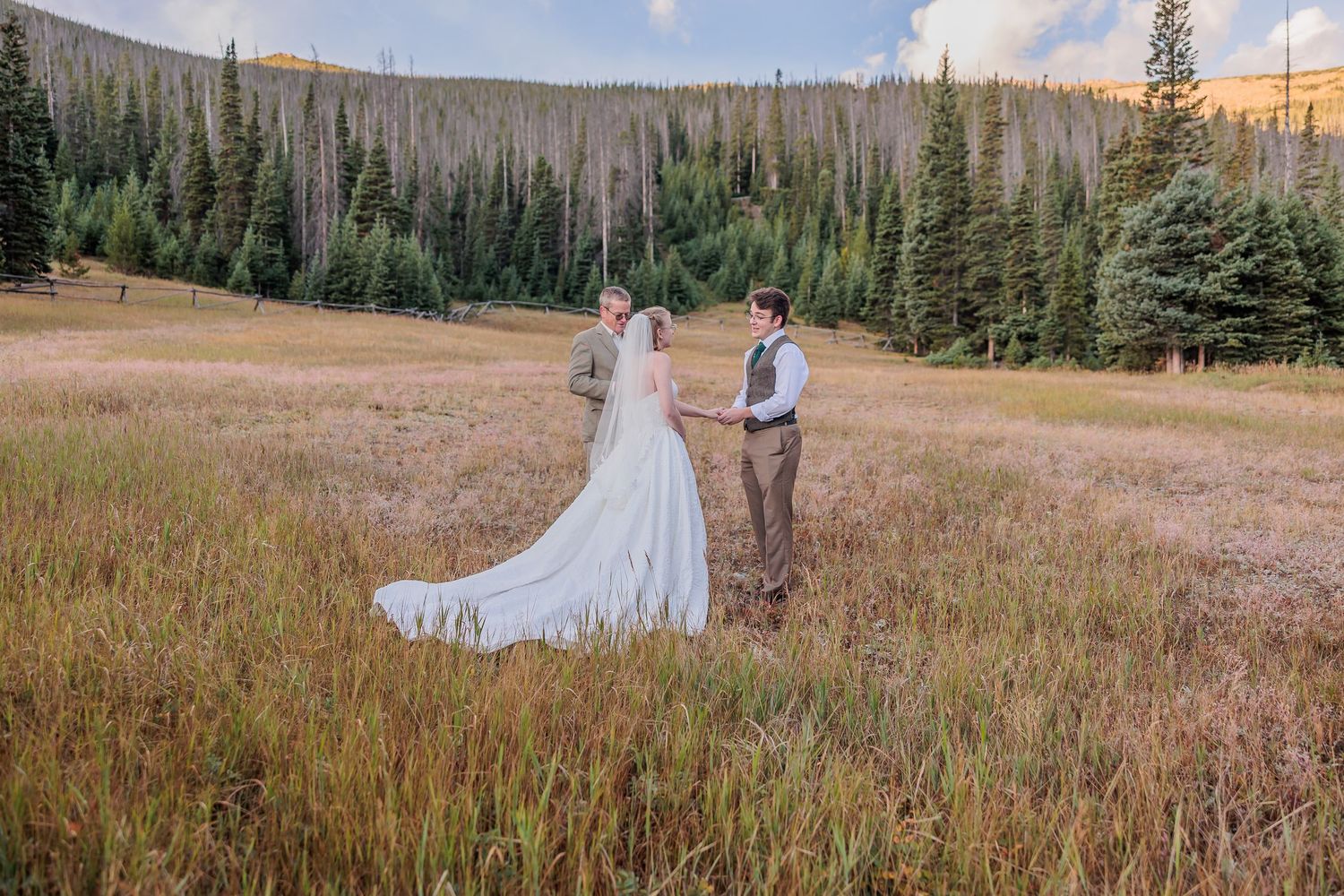 A long wedding dress train flows across a golden meadow with evergreen trees in the mountain landscape at sunset.