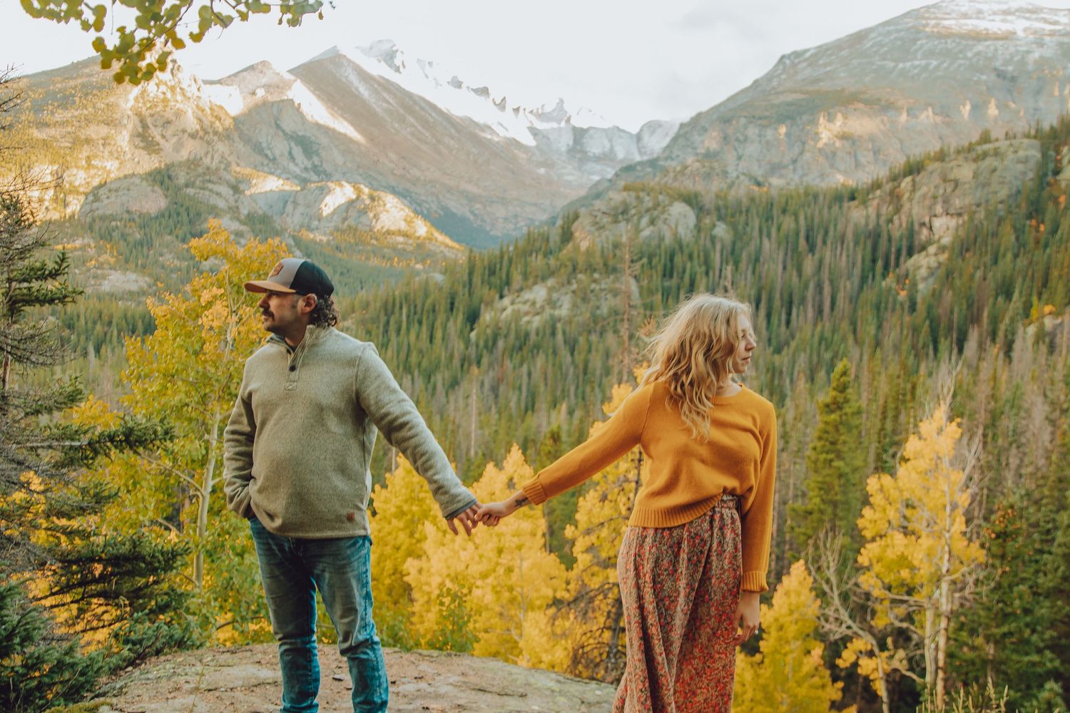 A couple walks through vibrant fall foliage in the Rocky Mountains with snow-capped peaks in the background.
