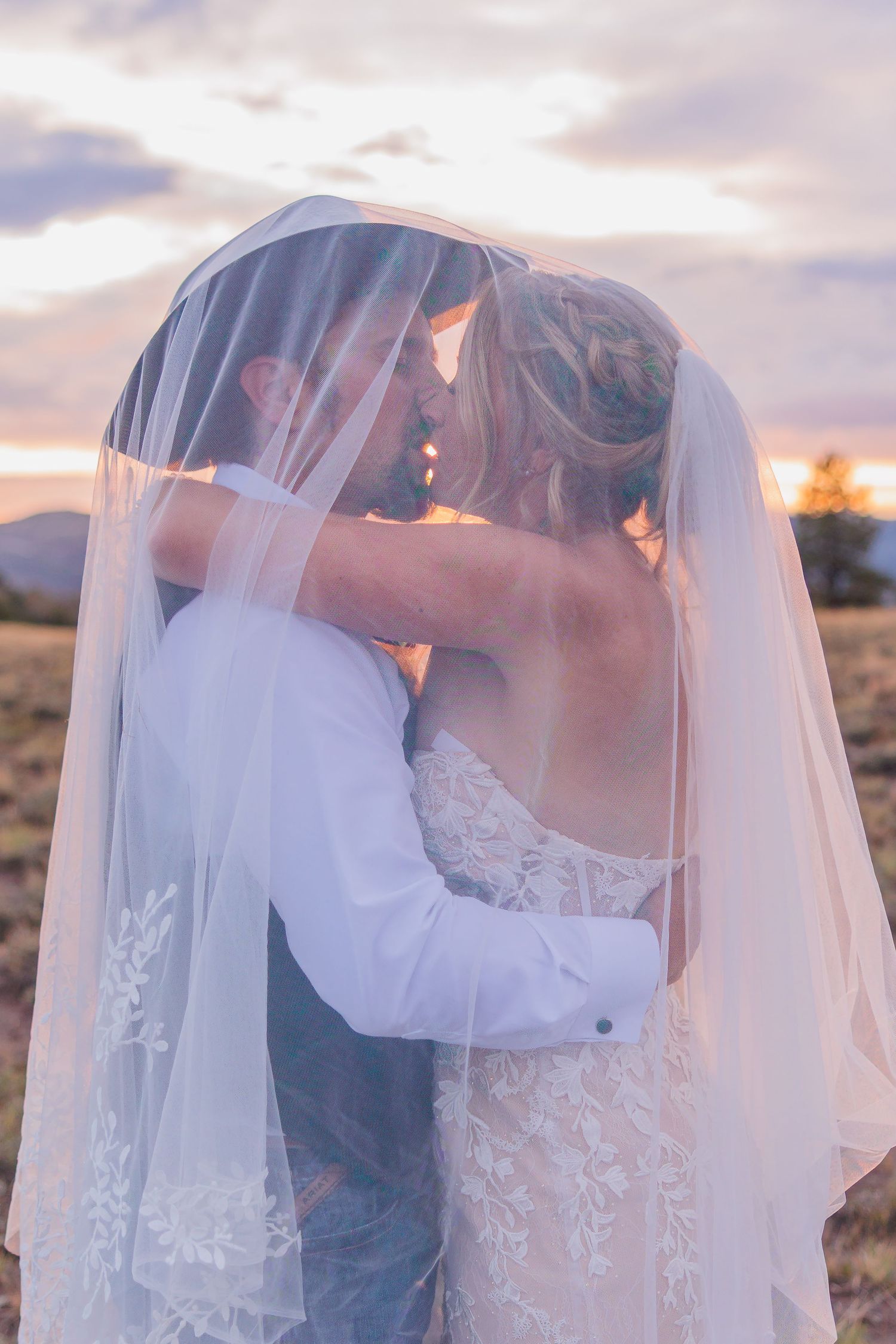 Romantic wedding couple embraces under flowing veil during golden sunset in desert landscape.