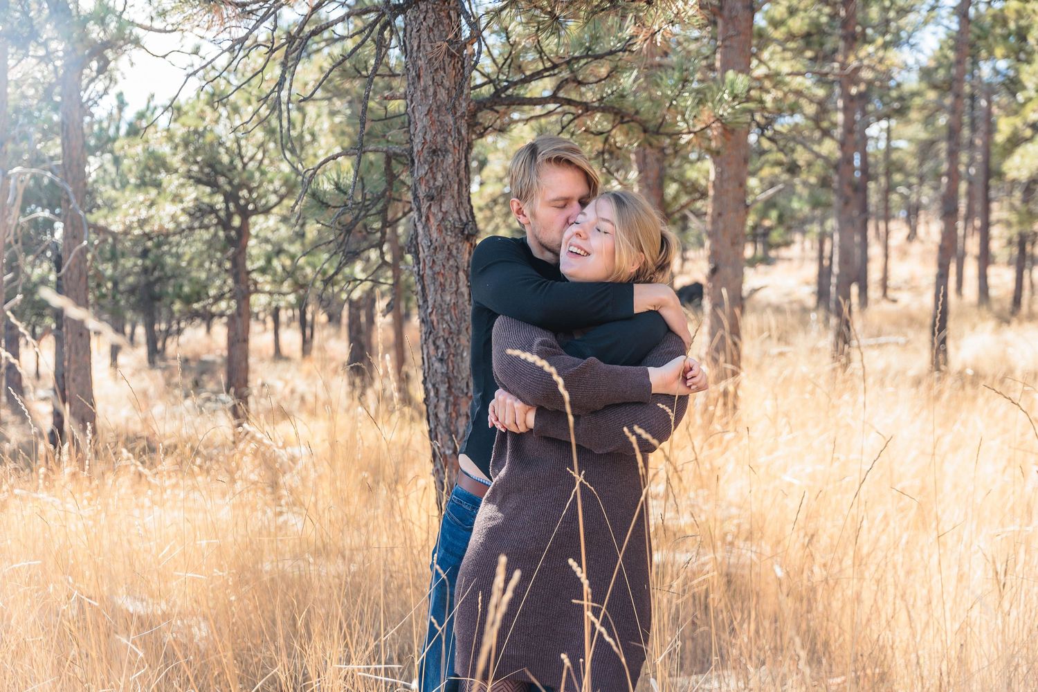 Two people embrace in a warm hug while standing in tall golden grass among pine trees in a forest setting.