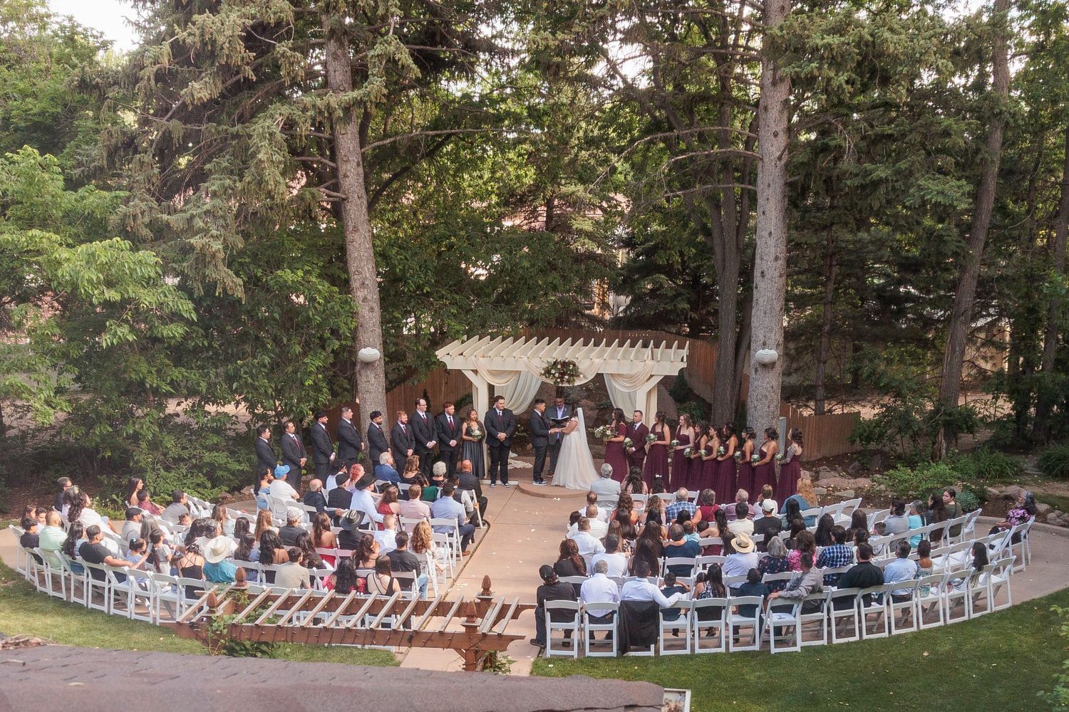 Panoramic view of an outdoor wedding ceremony setup in a wooded garden with white chairs and natural decor.