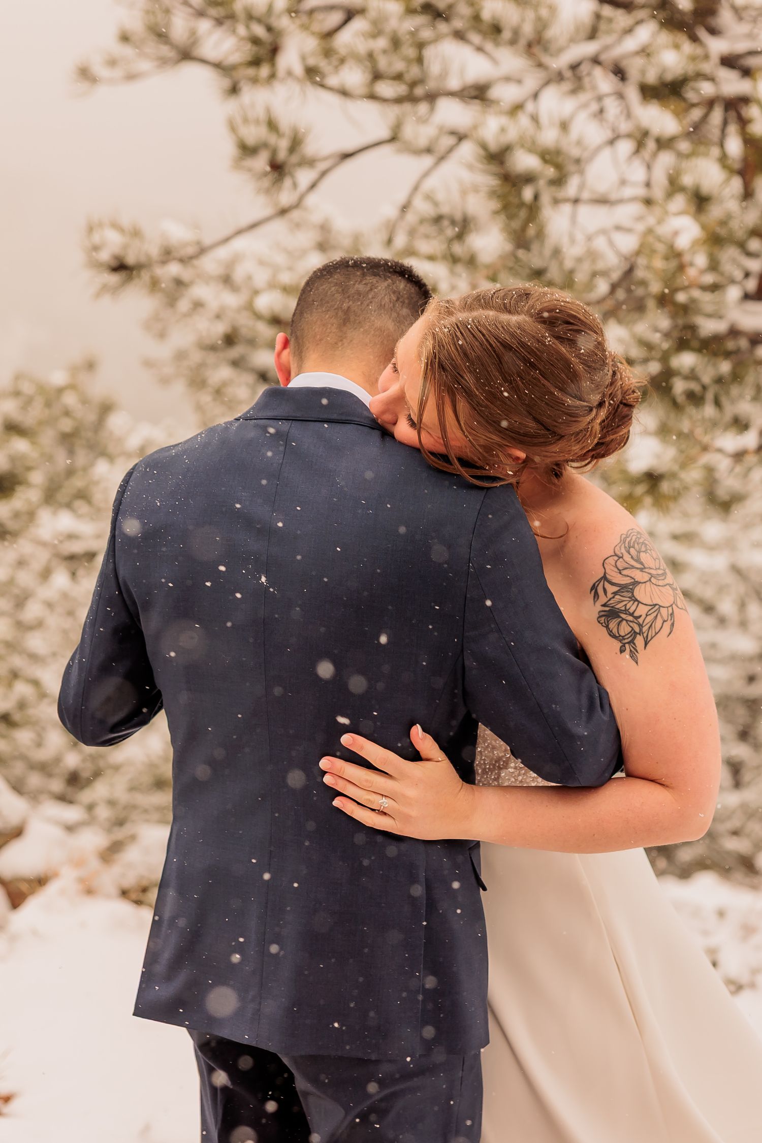 Bride and groom embrace in falling snow, bride showing floral tattoo in wedding dress, groom in navy suit.