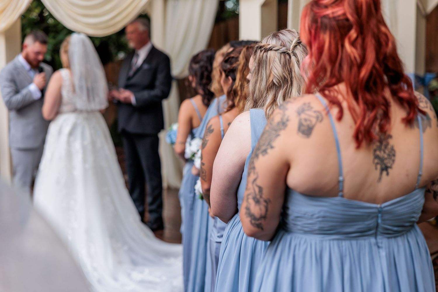 Bridesmaids in blue dresses with visible tattoos watch a wedding ceremony.